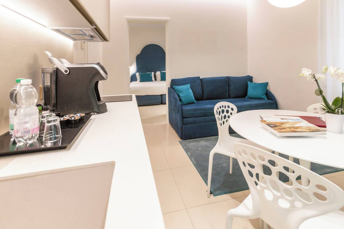 A modern hotel room with a kitchenette, a blue sofa with teal cushions, a white dining table with a white chair, and a potted orchid on the table.
