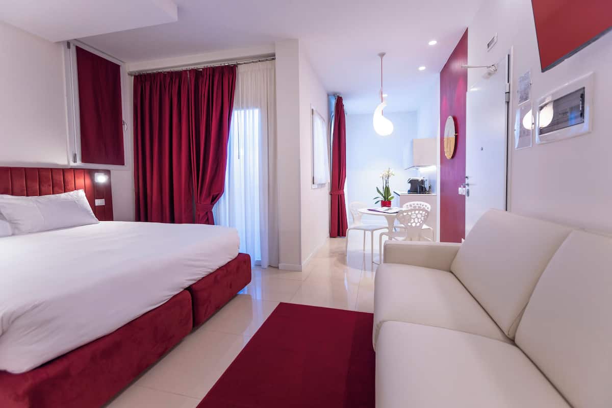 A modern hotel room with a large bed, a sofa, a small dining table, and a red accent wall.