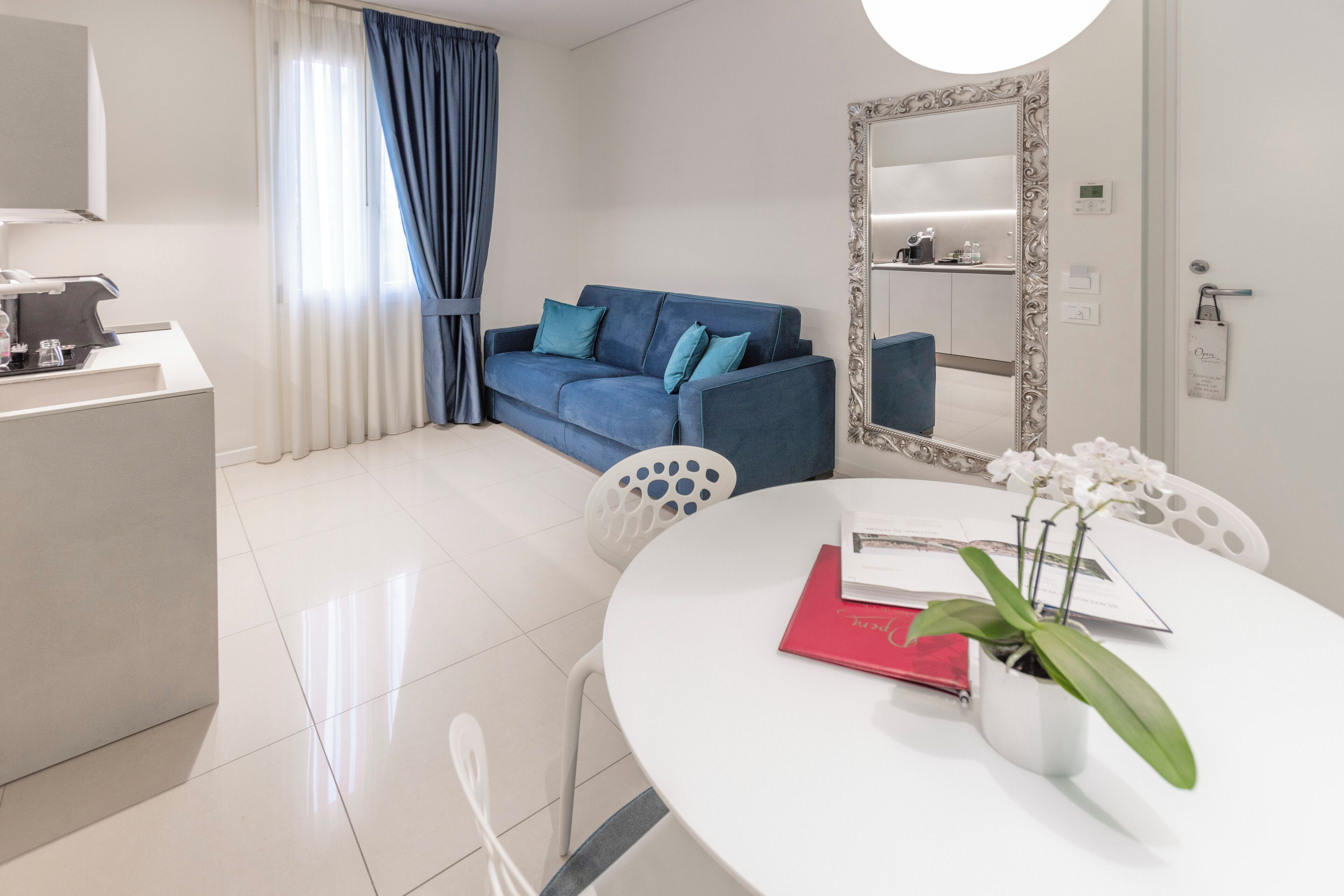 A modern hotel room with a white round table, a blue sofa, a white chair, a large mirror, and a potted orchid.