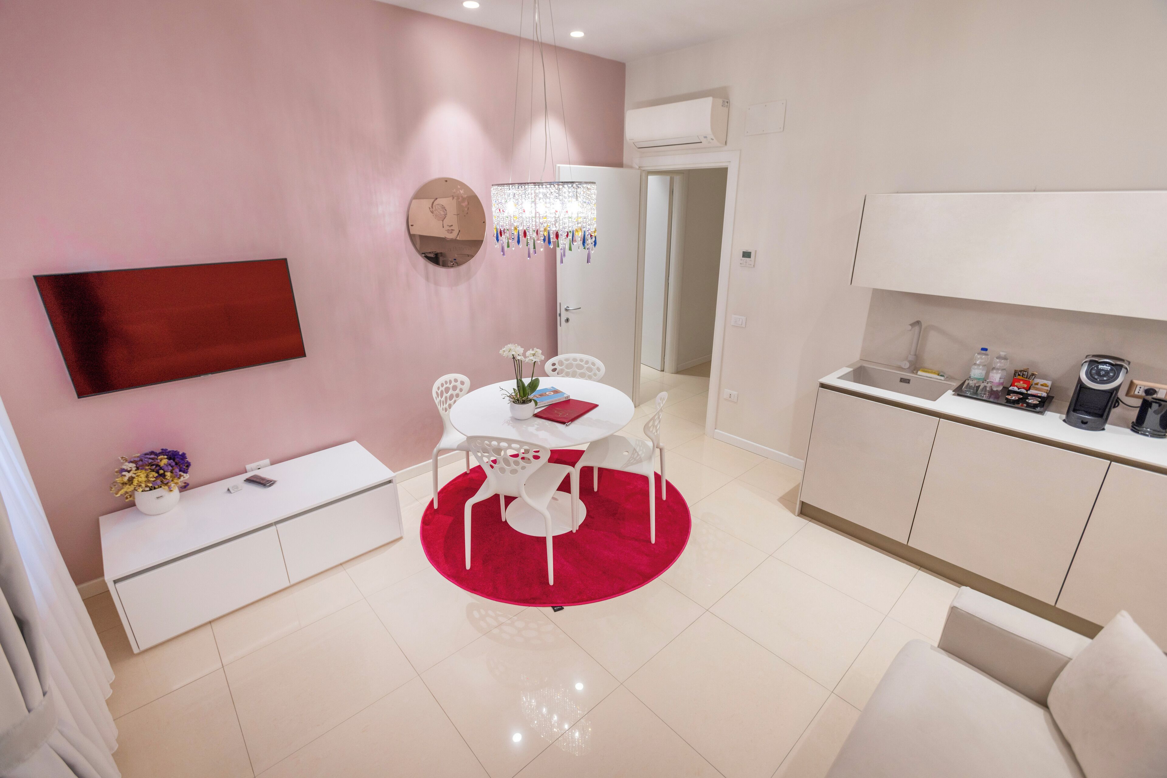 A modern kitchen with white cabinets and a countertop, a dining area with a white table and chairs, and a pink wall with a round mirror and a chandelier.