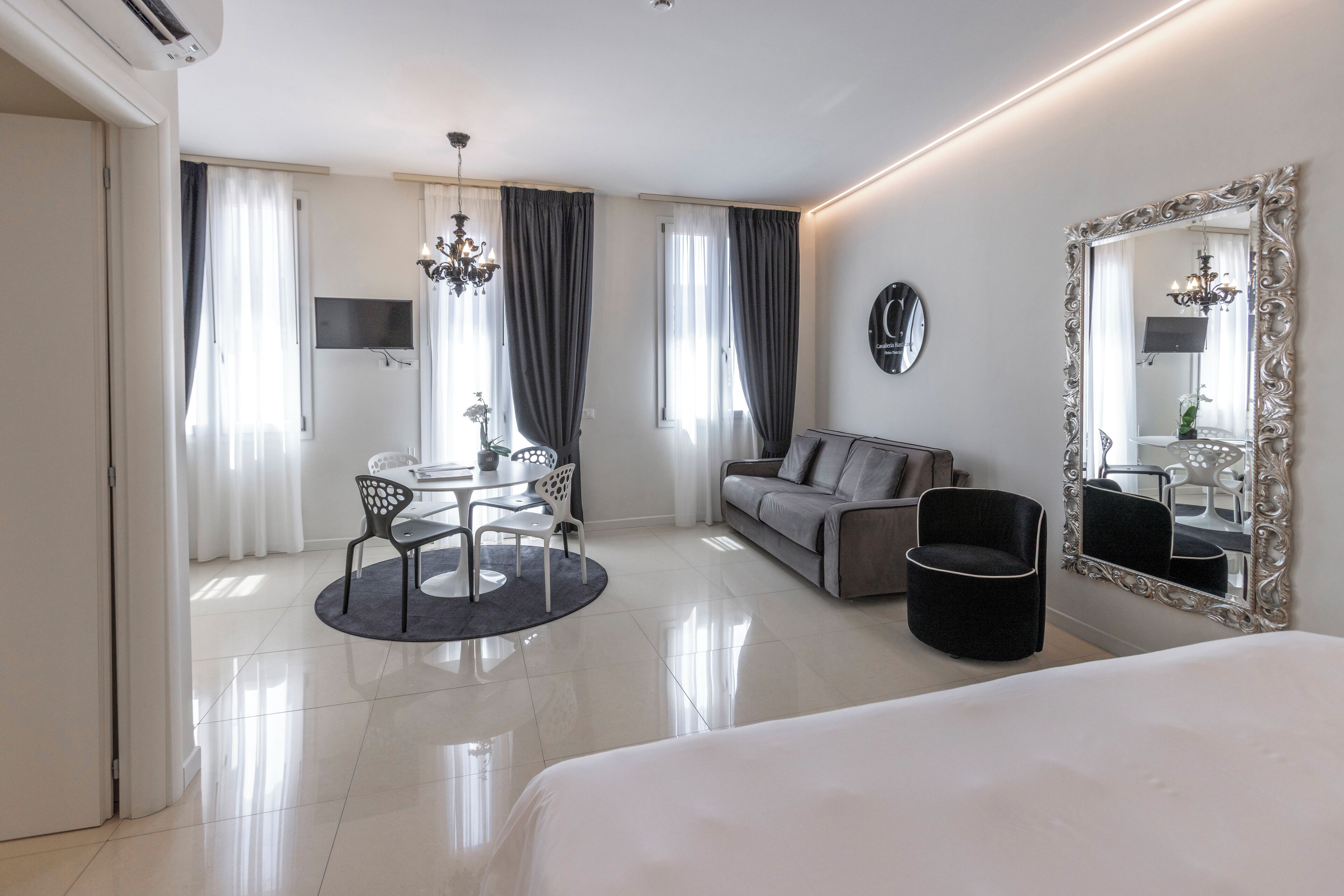 A modern hotel room with a bed, a sofa, a dining table with chairs, a chandelier, and a large mirror.
