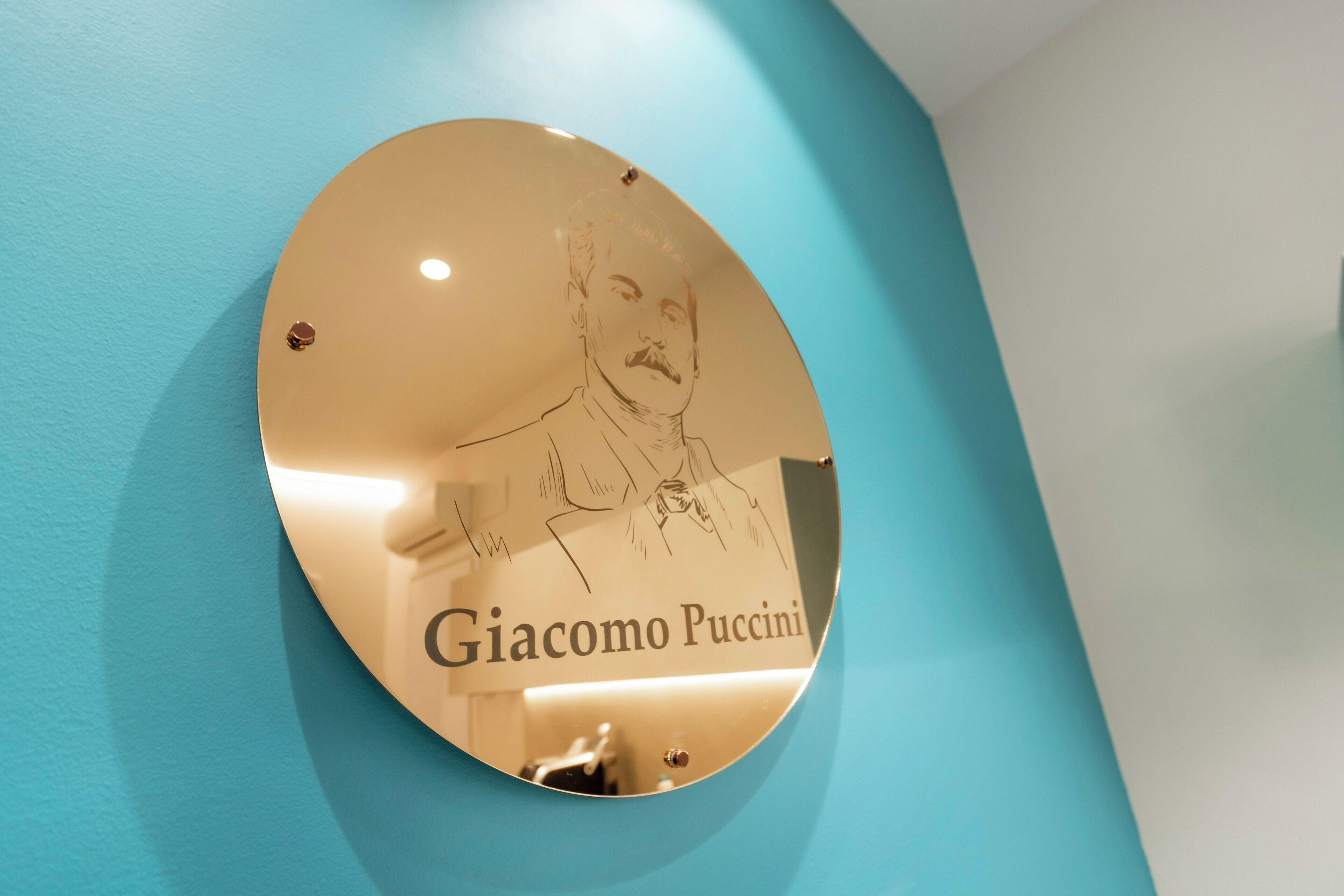 Una targa circolare con un ritratto e il nome Giacomo Puccini, fissata su una parete color acquamarina.