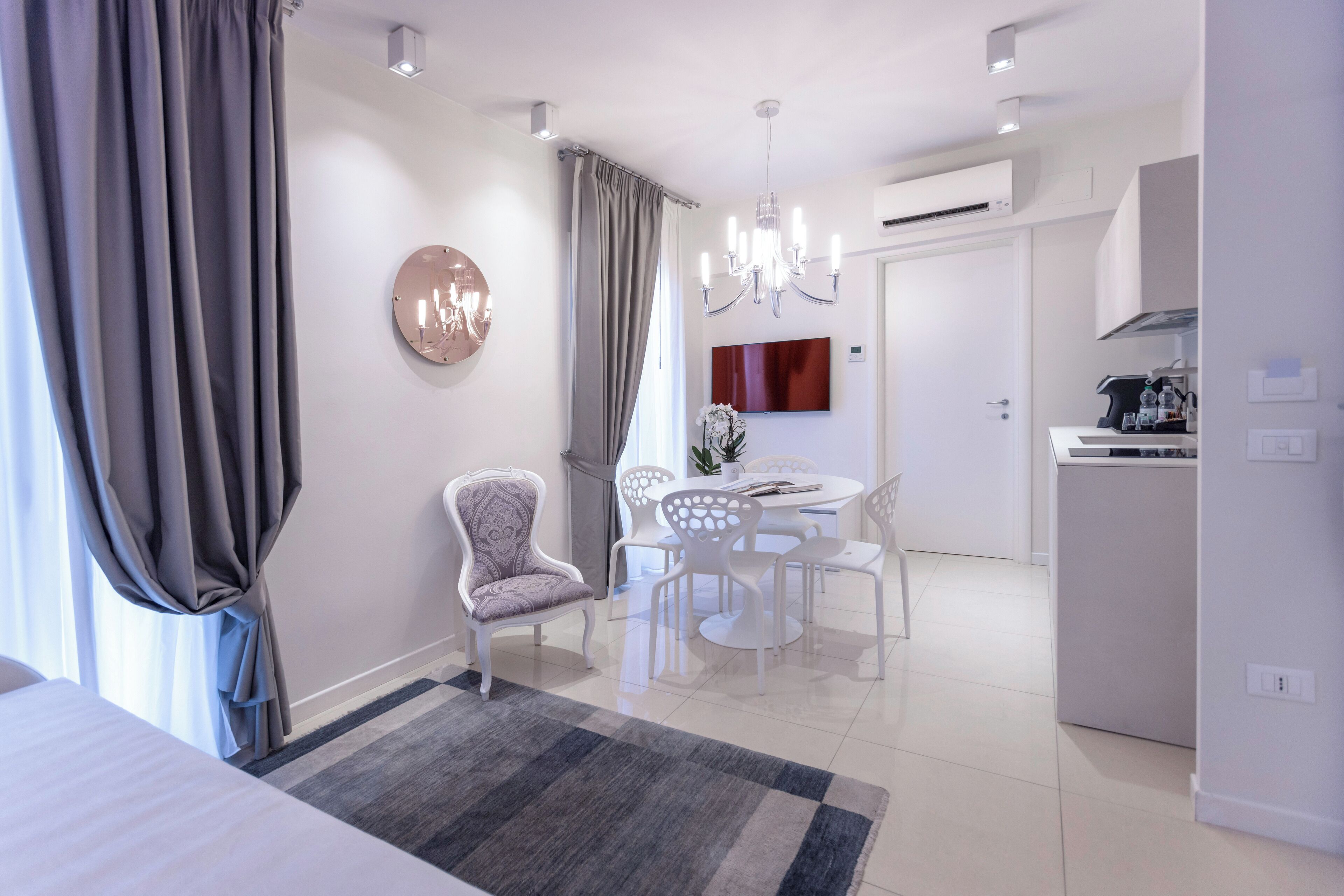 Een moderne hotelkamer met een bed, een zithoek met een tafel en stoelen, een kroonluchter en een grote spiegel aan de muur.