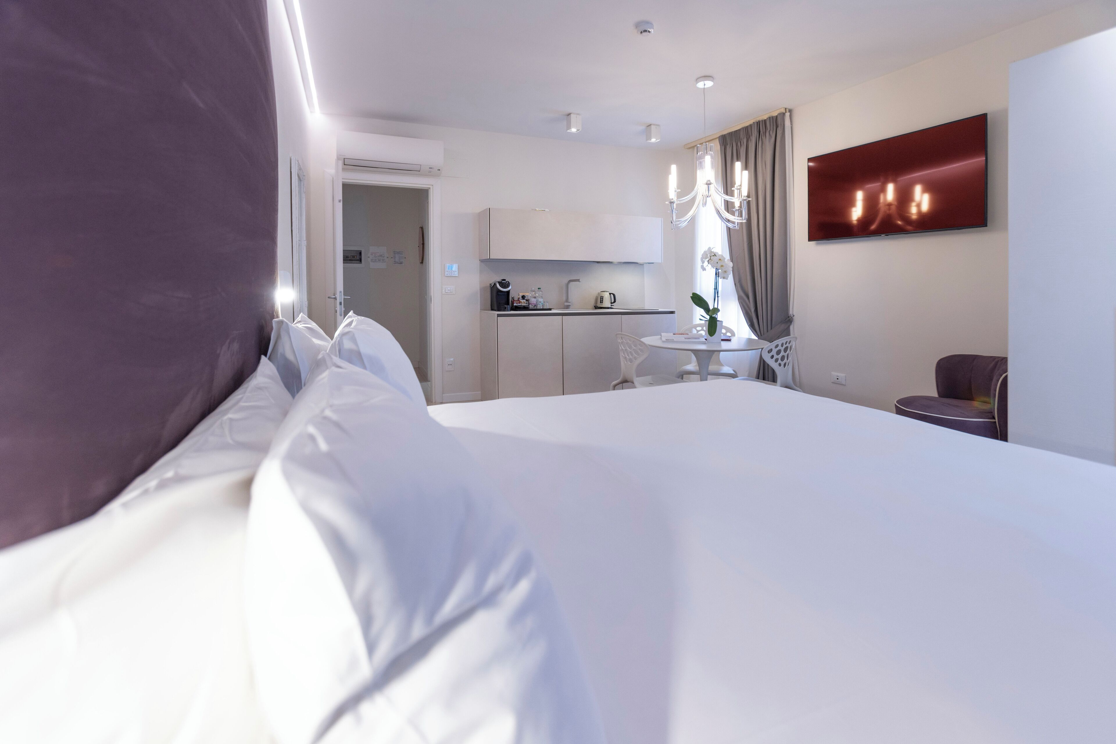 Una habitación de hotel moderna con una cama grande, una kitchenette pequeña, una zona de comedor y un televisor montado en la pared.