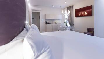 A modern hotel room with a large bed, a small kitchenette, a dining area, and a TV mounted on the wall.
