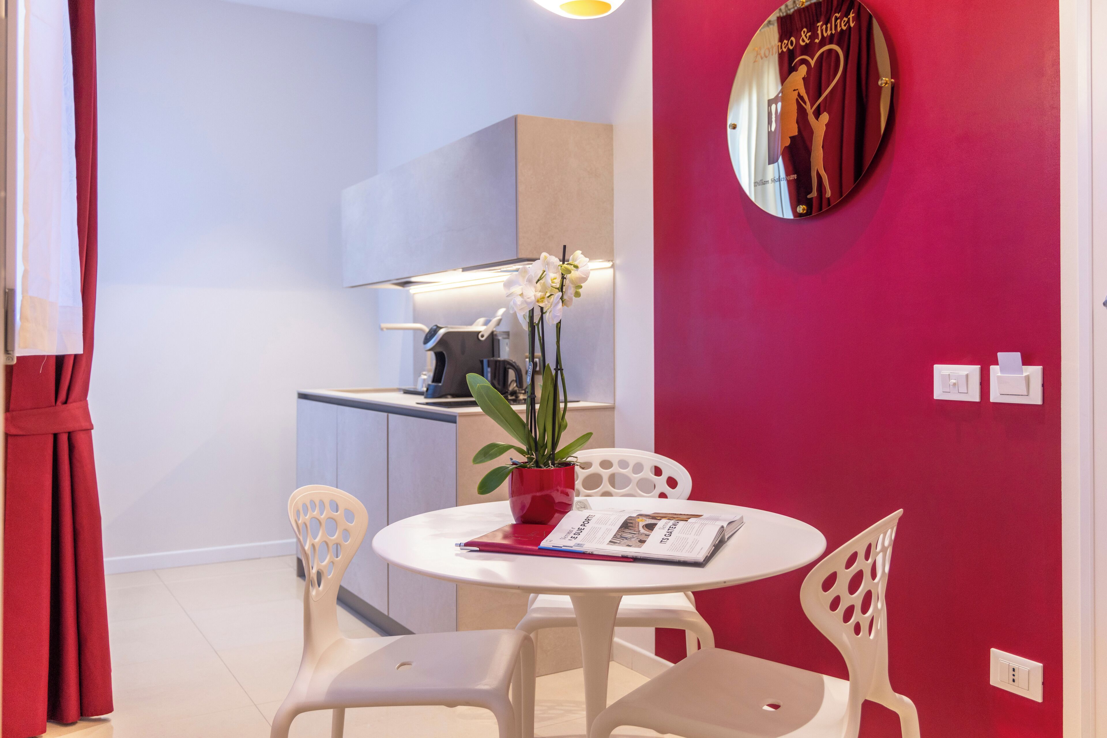 A small dining area with a white table and chairs, a red potted plant, and a wall clock with a heart design.