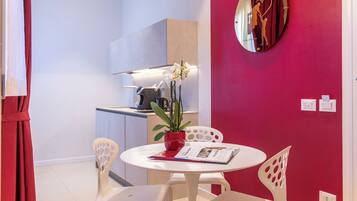 A small dining area with a white table and chairs, a red potted plant, and a wall clock with a heart design.