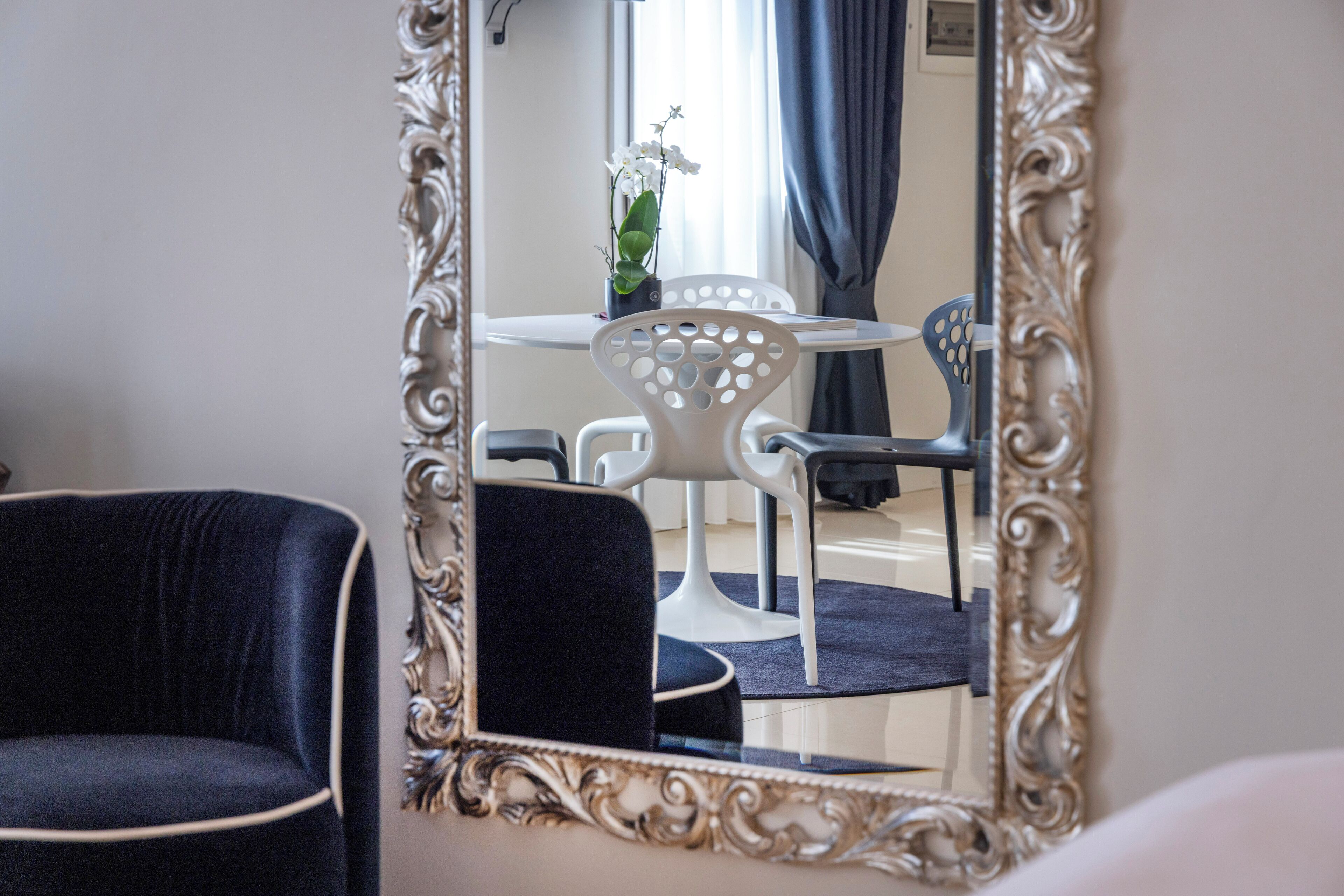 A room with a large ornate mirror, a dark upholstered chair, a white table with a potted orchid, and a blue chair.