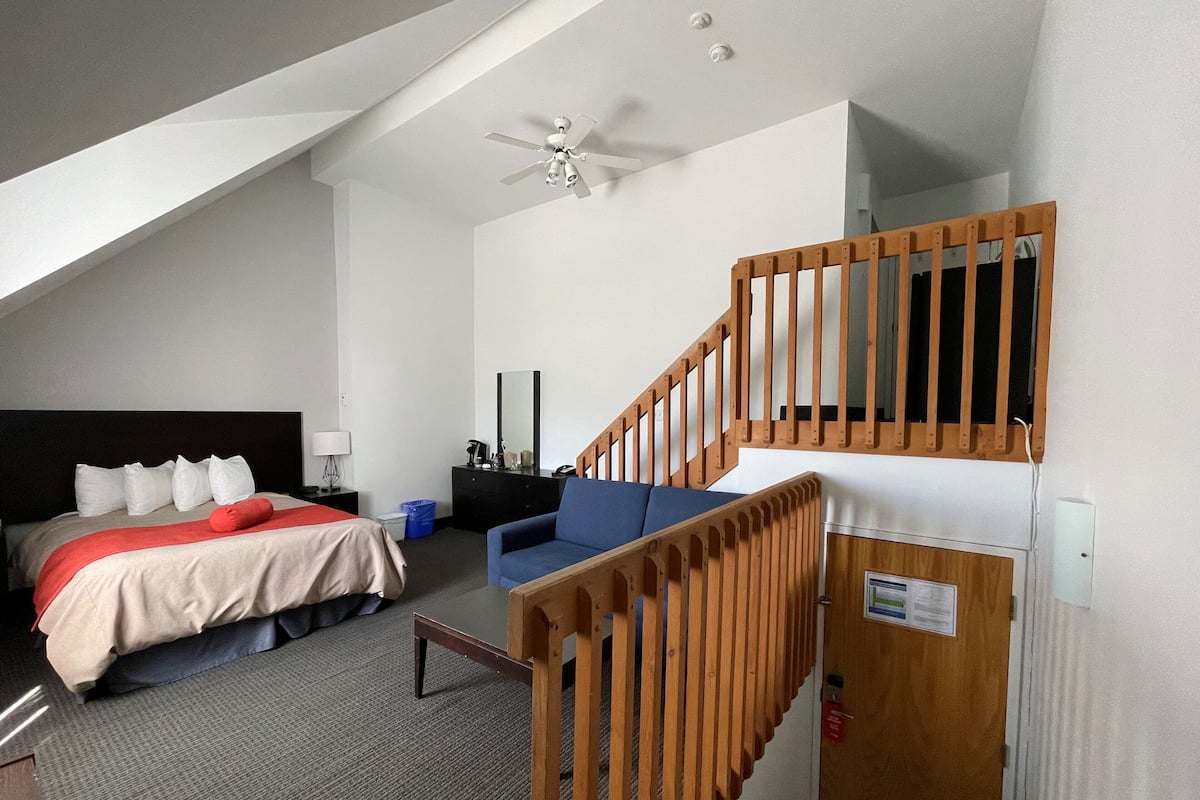 Een hotelkamer met een bed, een bureau, een stoel, een houten trap en een plafondventilator.