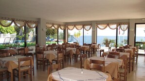 A dining area with tables and chairs, large windows with curtains, and a view of the sea. - Karina (Corfu)