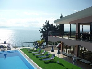 A poolside area with lounge chairs and umbrellas, overlooking a body of water and a mountainous landscape. - Karina (Corfu)