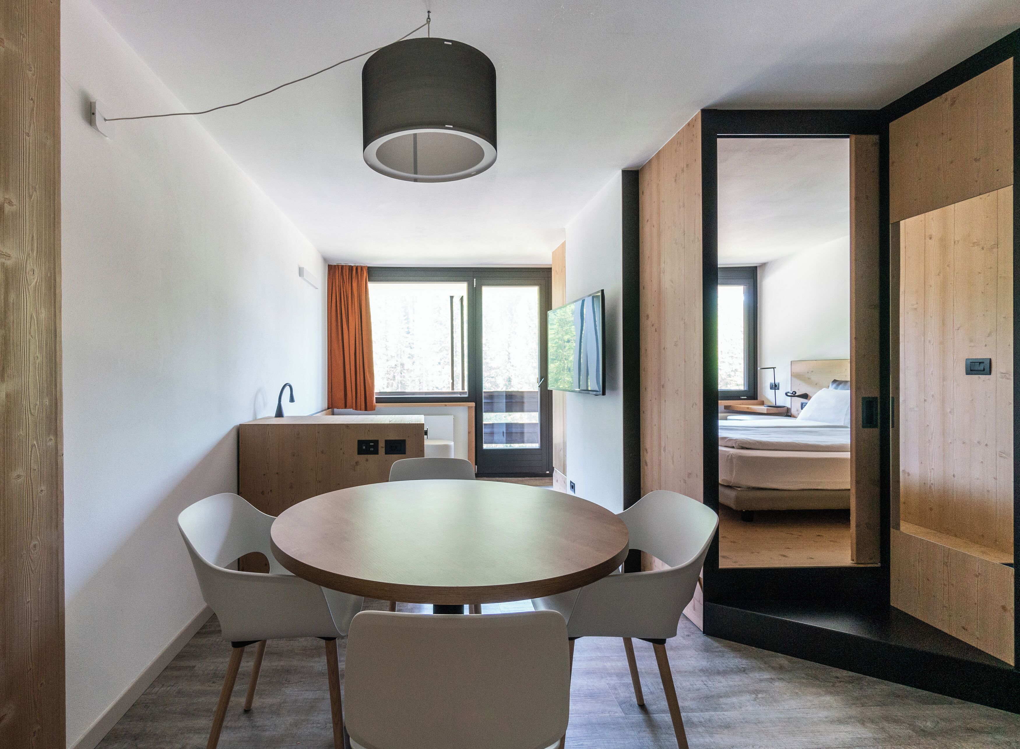Una habitación de hotel moderna con una mesa redonda, sillas blancas, una pared de acento de madera, un televisor y una cama con cabecera de madera.