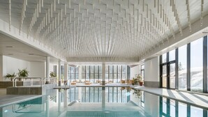 A large indoor swimming pool with clear glass walls, a modern ceiling design, and seating areas with views of the outdoors. - Ambiez Residence (Pinzolo)