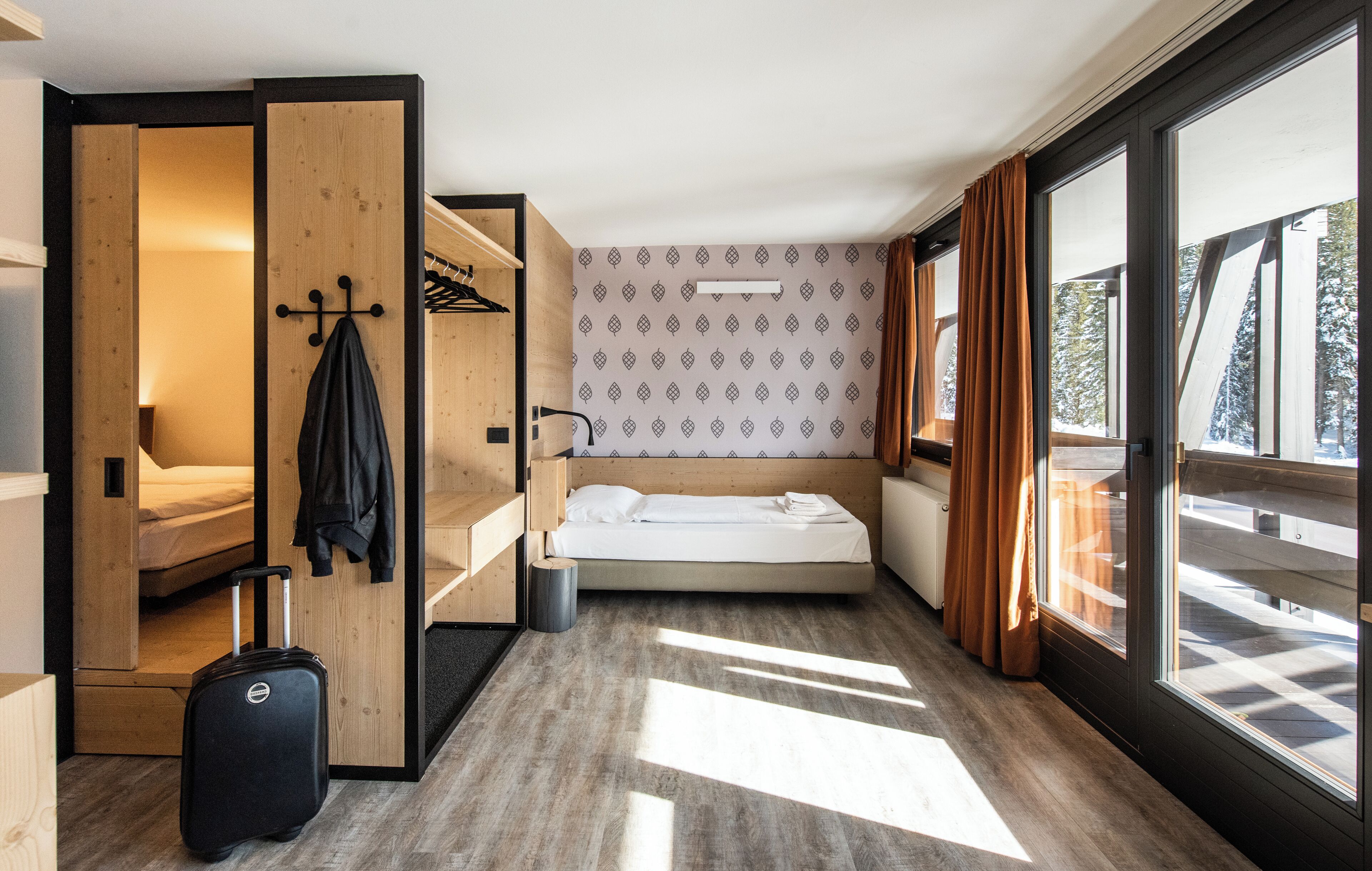 A compact hotel room with a bed, a small seating area, a suitcase, and a view through large windows.