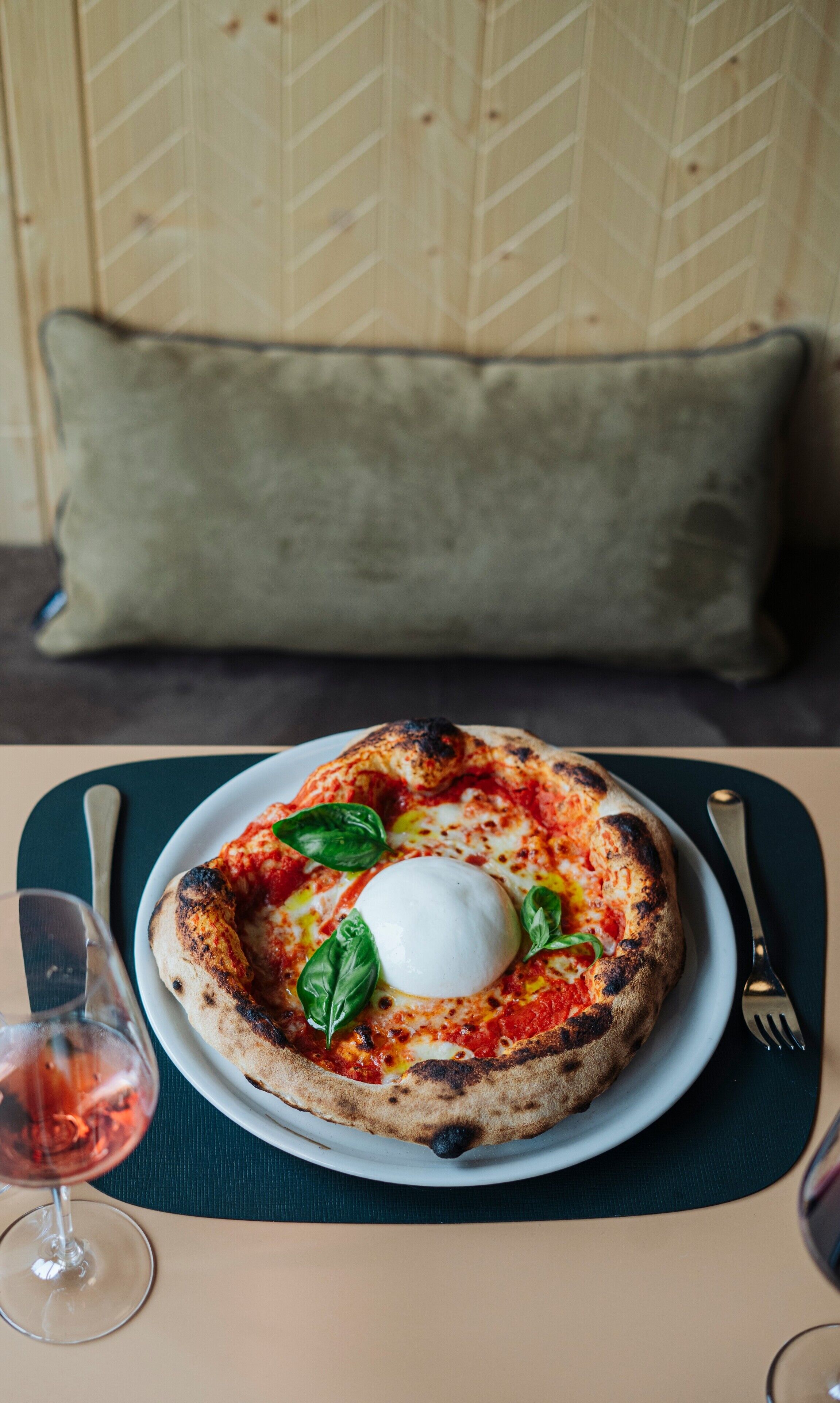 Uma pizza com ovo escaldado e manjericão em um prato, acompanhada por um copo de vinho tinto, sobre uma mesa com sousplat.