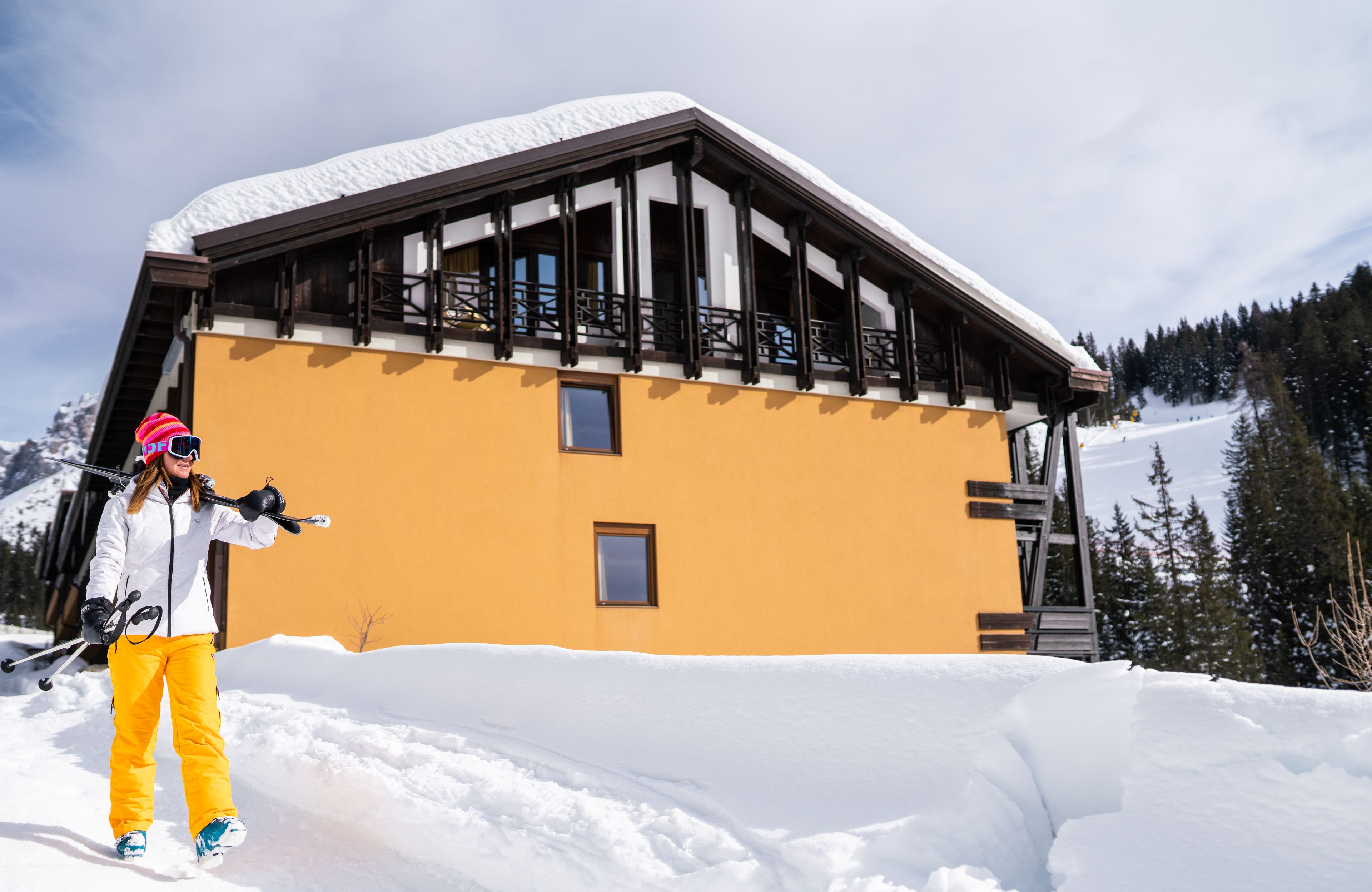 A ski resort building with a yellow facade and wooden balcony, surrounded by snow-covered mountains.