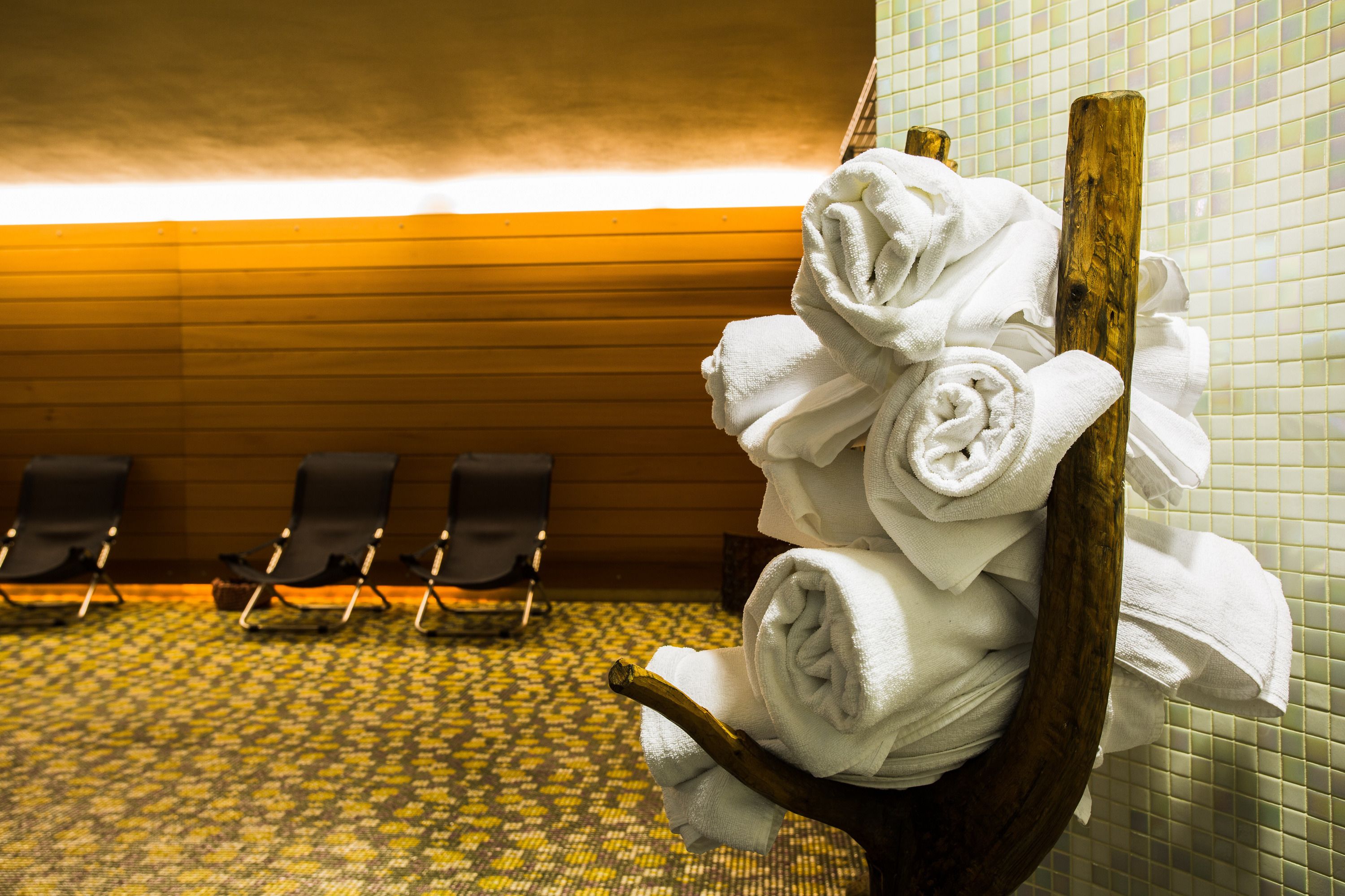 A bathroom with a wooden towel rack holding neatly folded towels, a patterned floor, and a row of black chairs in the background.