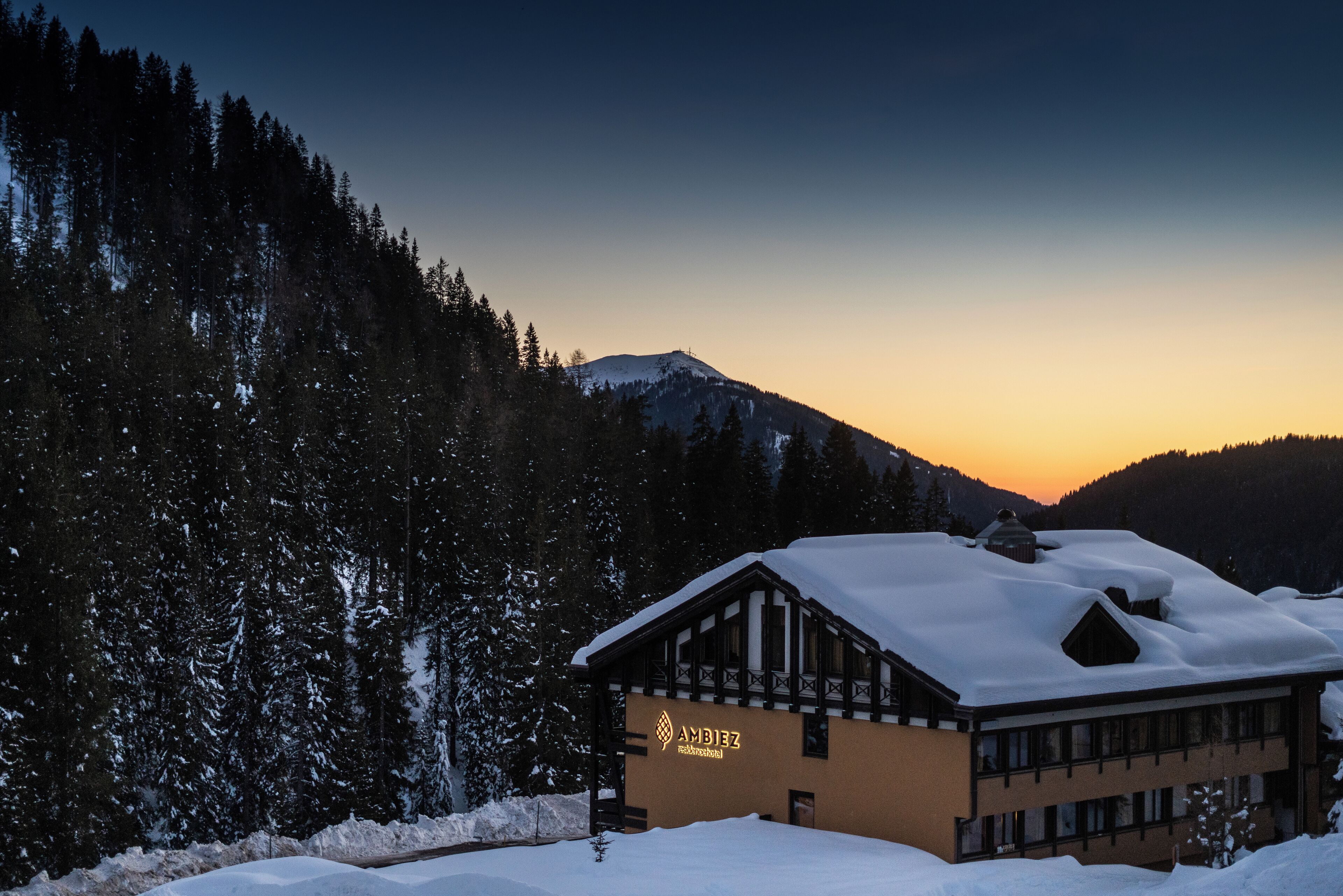 A lodge with a snow-covered roof, surrounded by a forest and mountains.