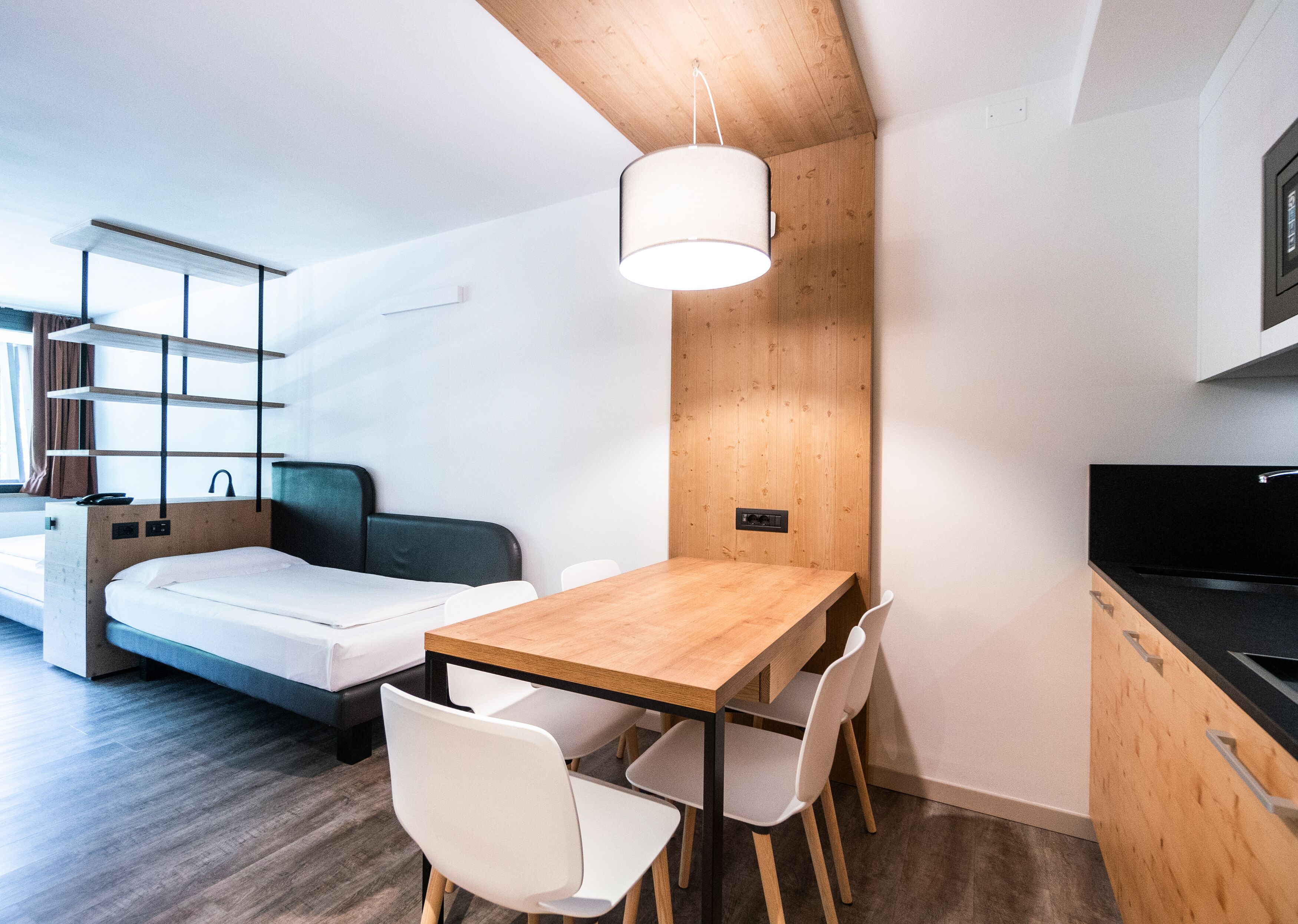 A compact room with a bed, a wooden dining table with chairs, a kitchen area with a black countertop and sink, and a hanging light fixture.