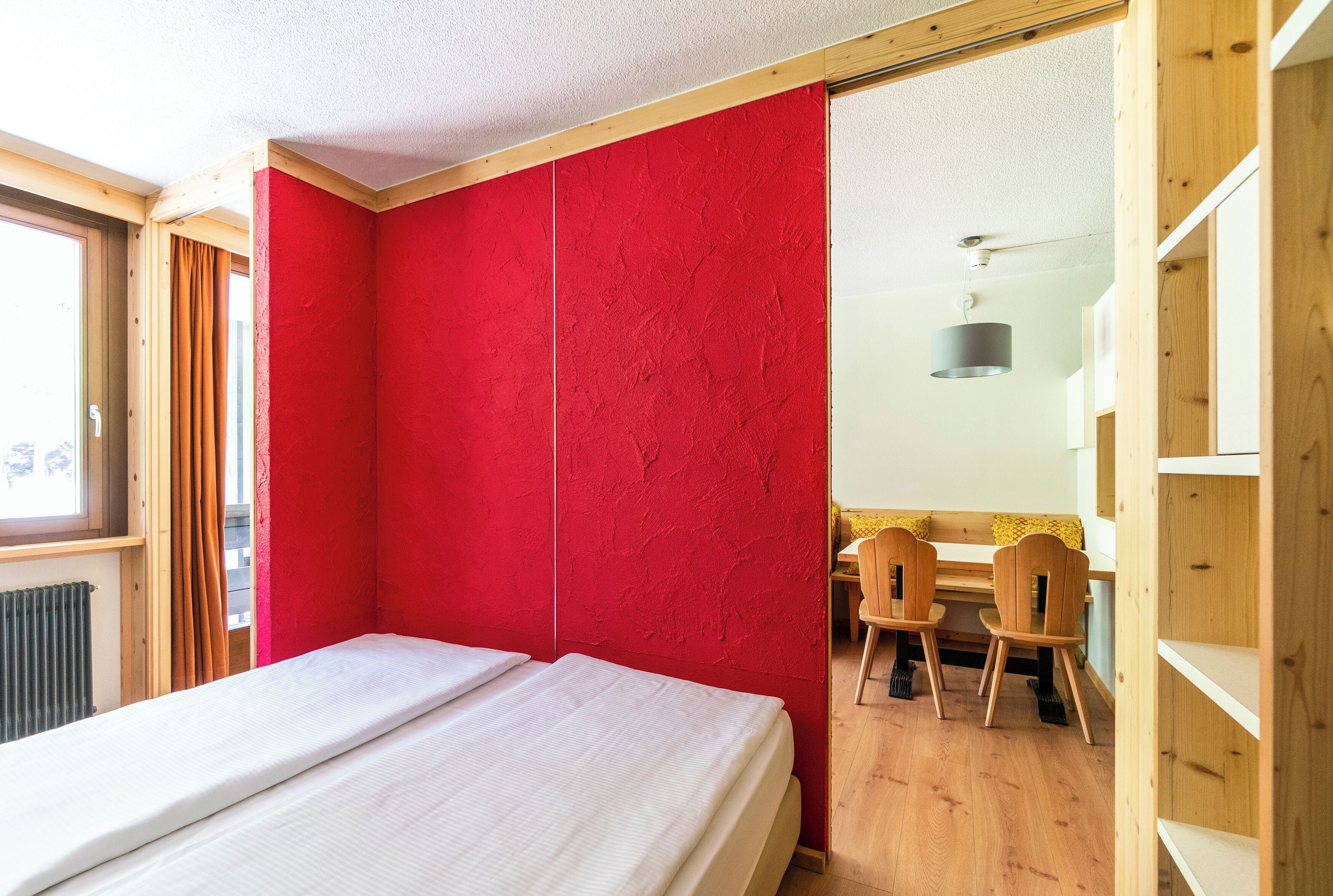 A bedroom with a bed, a red wall, a wooden floor, and a desk with chairs.