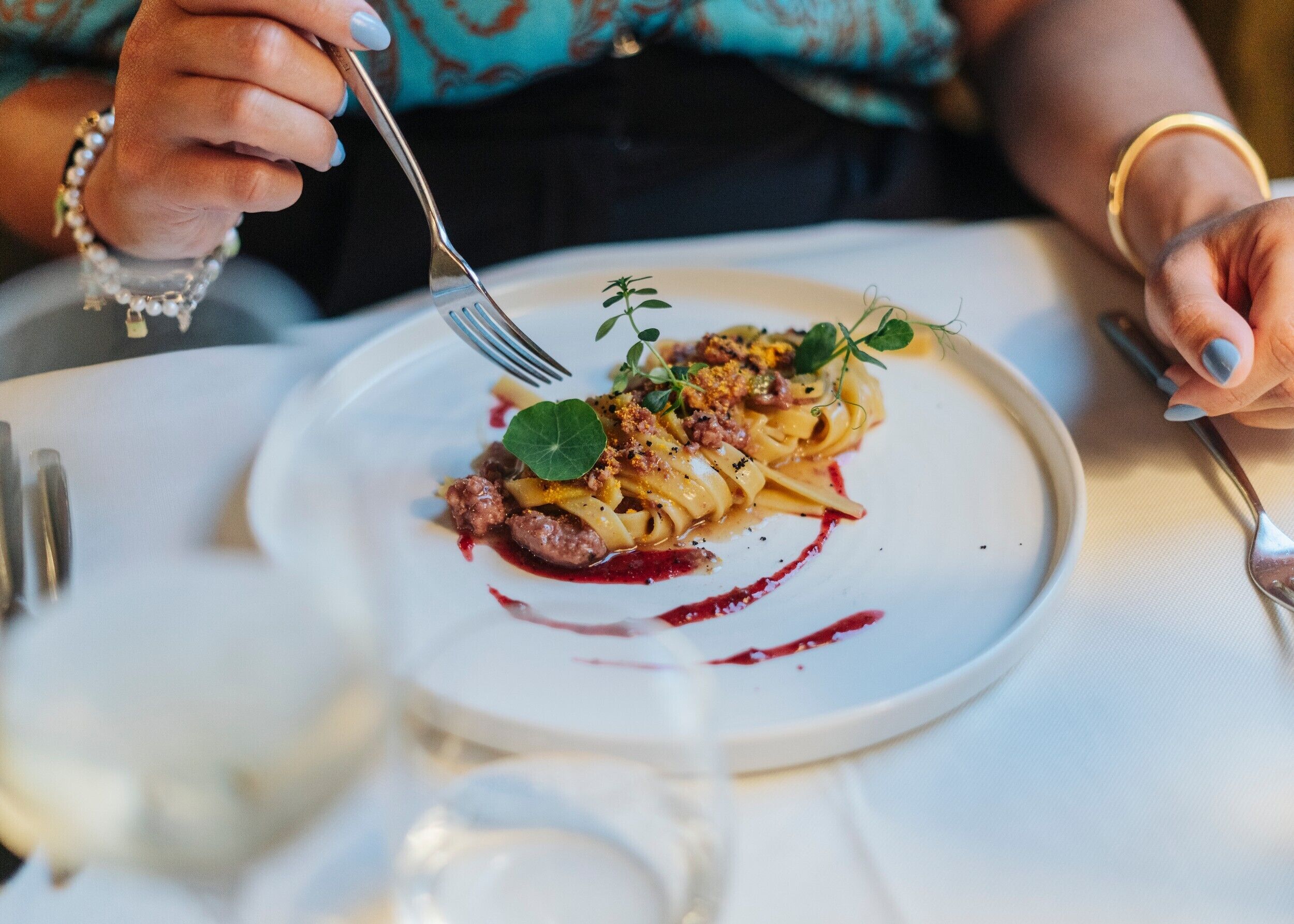 Une personne est assise à une table, devant une assiette de pâtes, avec une fourchette et un verre d’eau.