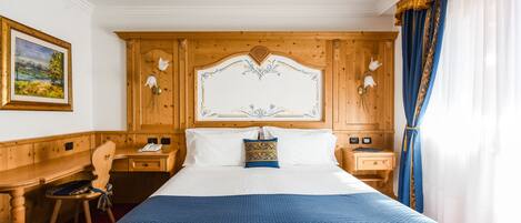 A hotel room with a wooden headboard, a bed with a blue patterned bedspread, a desk with a chair, and a painting on the wall.