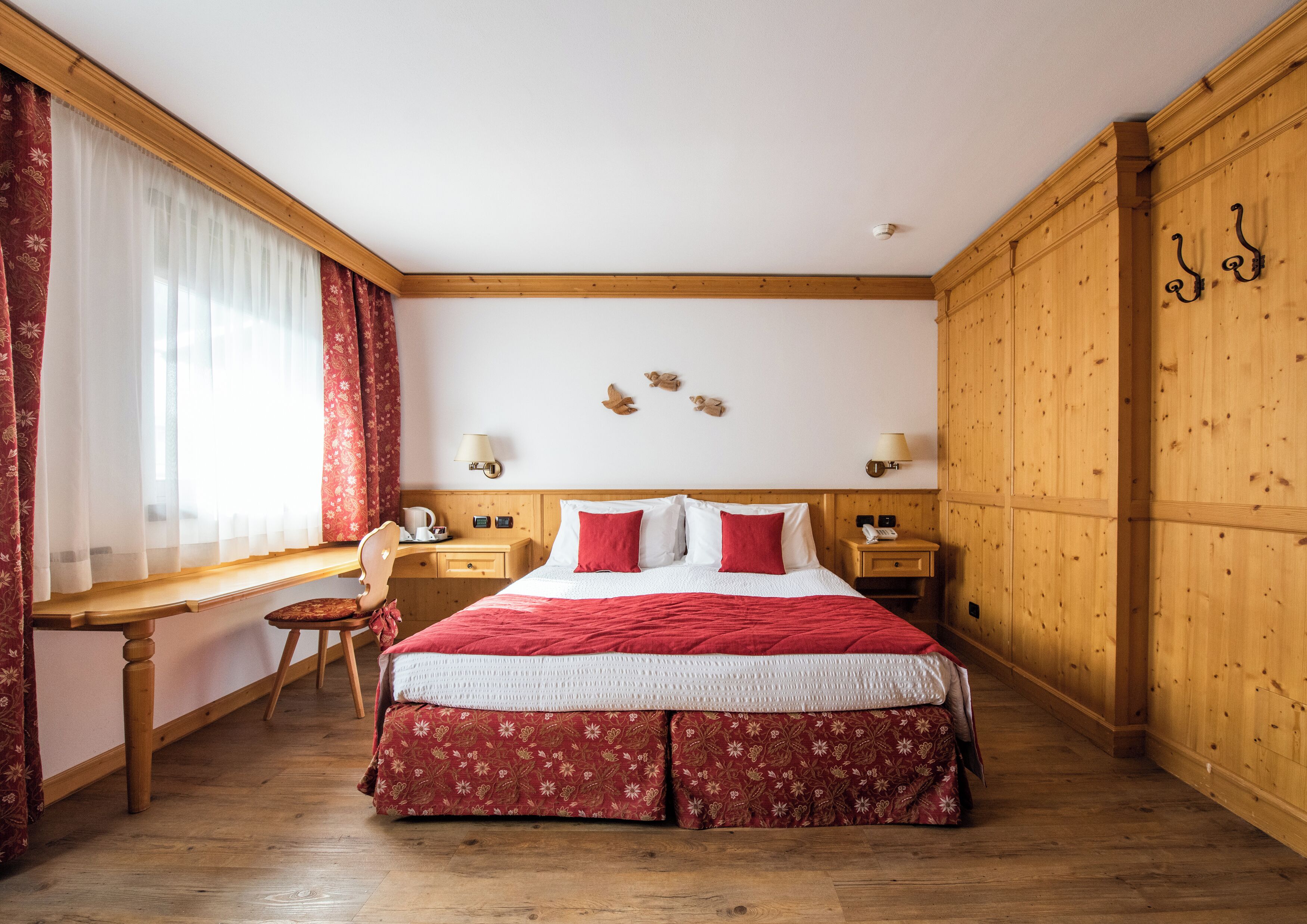 Habitación de hotel con piso de madera, una cama grande con ropa de cama roja, un escritorio con silla, dos mesitas de noche con lámparas y una ventana con cortinas rojas.