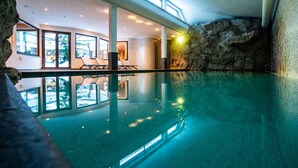 Ein Hallenbad mit klarem Wasser, Steinwänden und einem Sitzbereich mit Stühlen und Tischen.