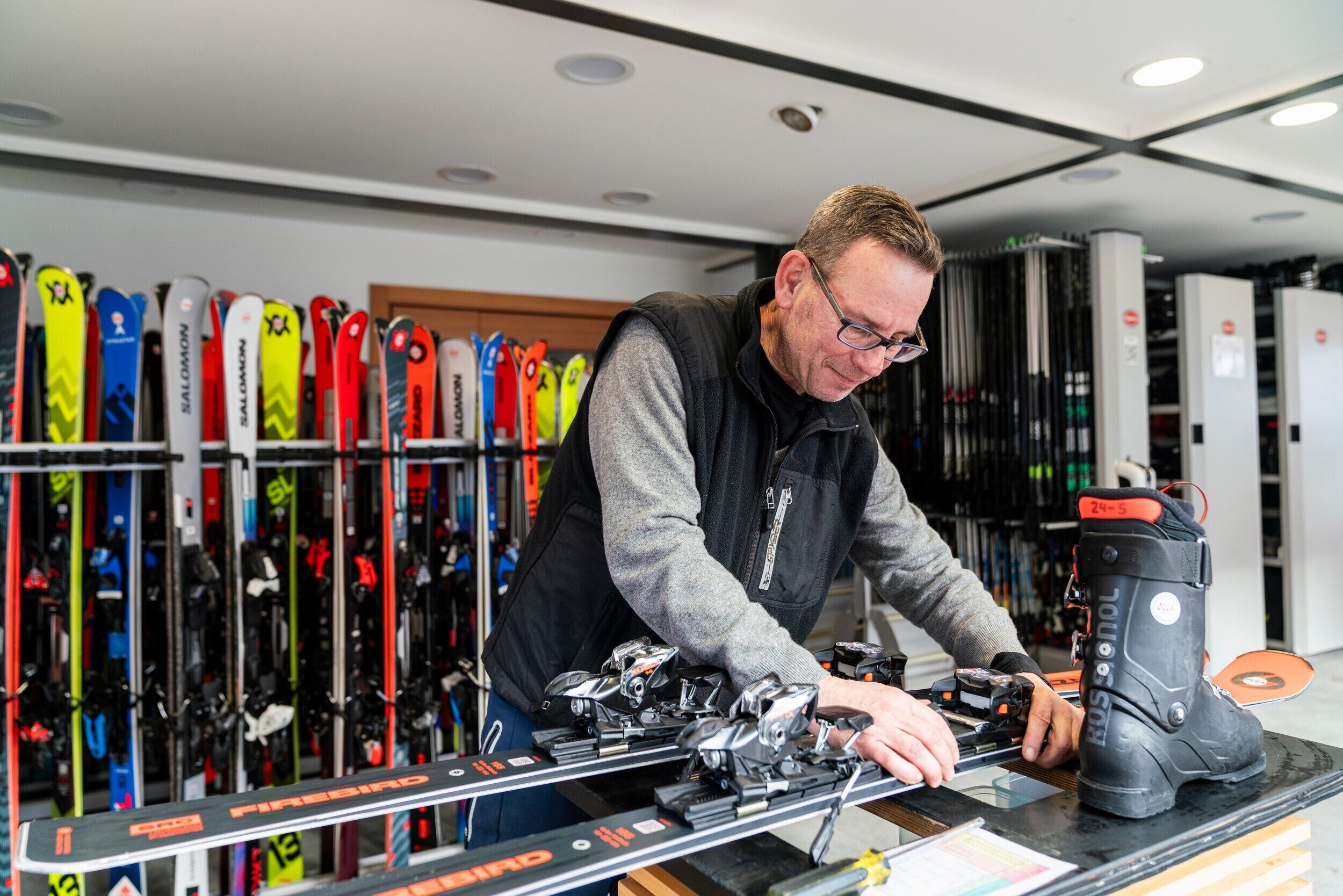 A person adjusting ski bindings at a counter with skis displayed in the background.