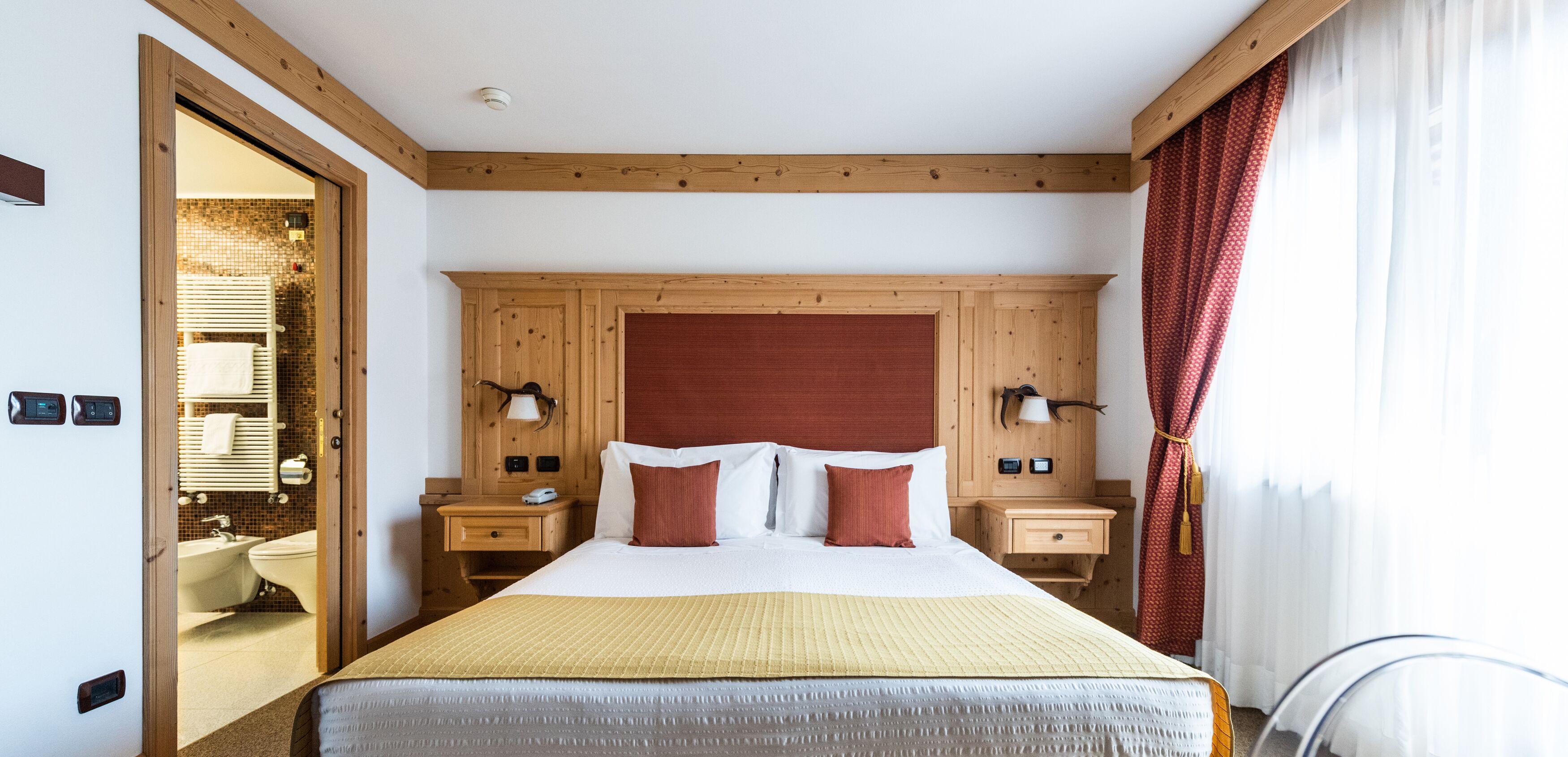 A hotel room with a wooden headboard, two bedside tables with lamps, a bed with white and red pillows, and a bathroom visible through an open door.