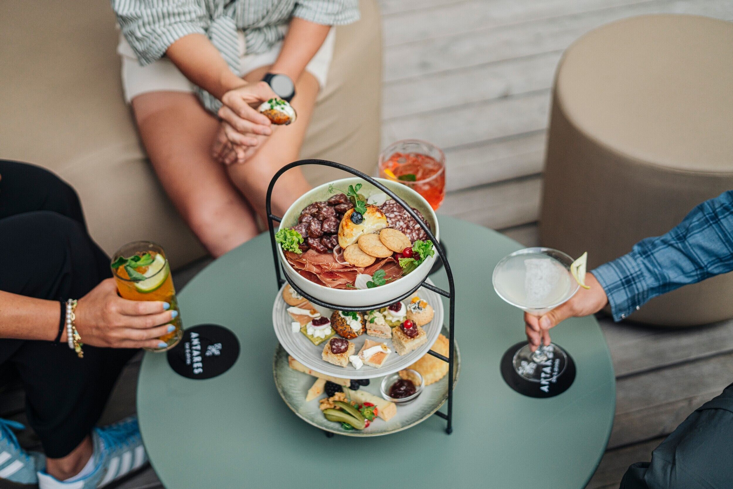 Bandeja de tres niveles con diferentes aperitivos y una bebida sobre una mesa redonda.