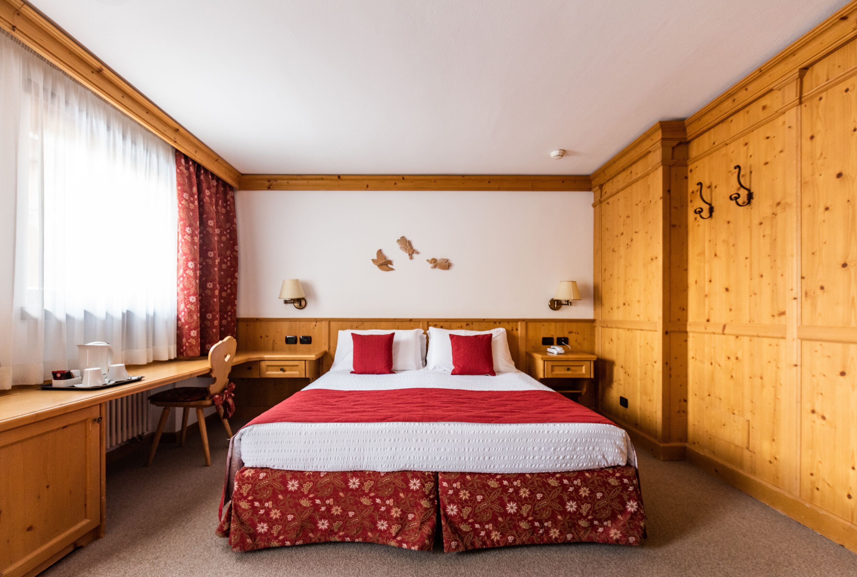A hotel room with a large bed, wooden headboard, two bedside tables with lamps, a desk with a chair, and a window with curtains.