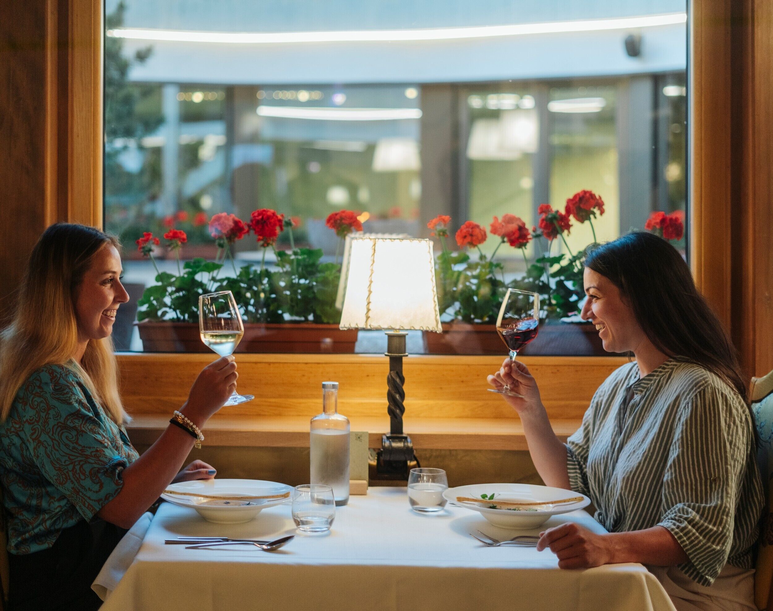 Dos personas cenando en una mesa con una lámpara, una botella y vistas florales por la ventana.