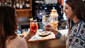Deux personnes, chacune tenant un verre, sont debout au bar, devant un comptoir en bois. On aperçoit, en arrière-plan, des étagères remplies de bouteilles.