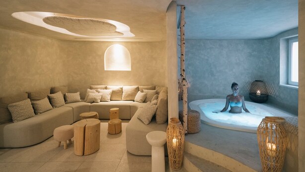 Une salle de spa moderne avec un grand canapé incurvé, un petit coin salon avec des tabourets et un jacuzzi dans lequel on aperçoit une personne à l’arrière-plan.