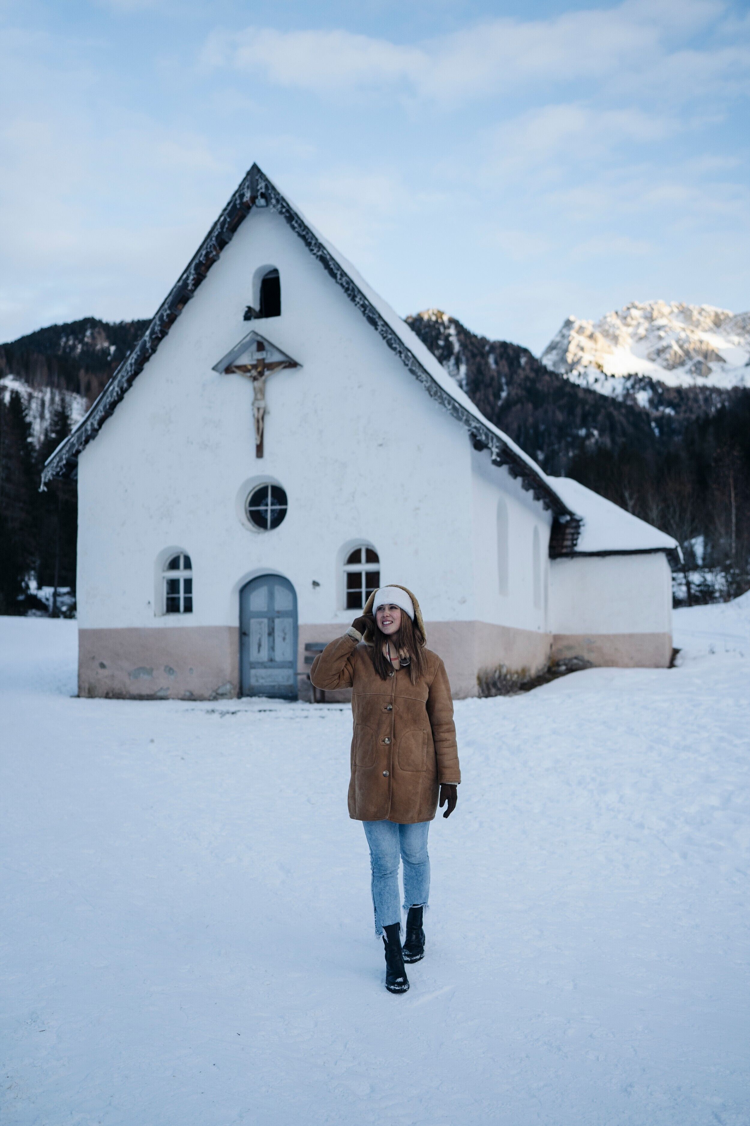 A person standing in front of a white building with a triangular roof, surrounded by snow and mountains.