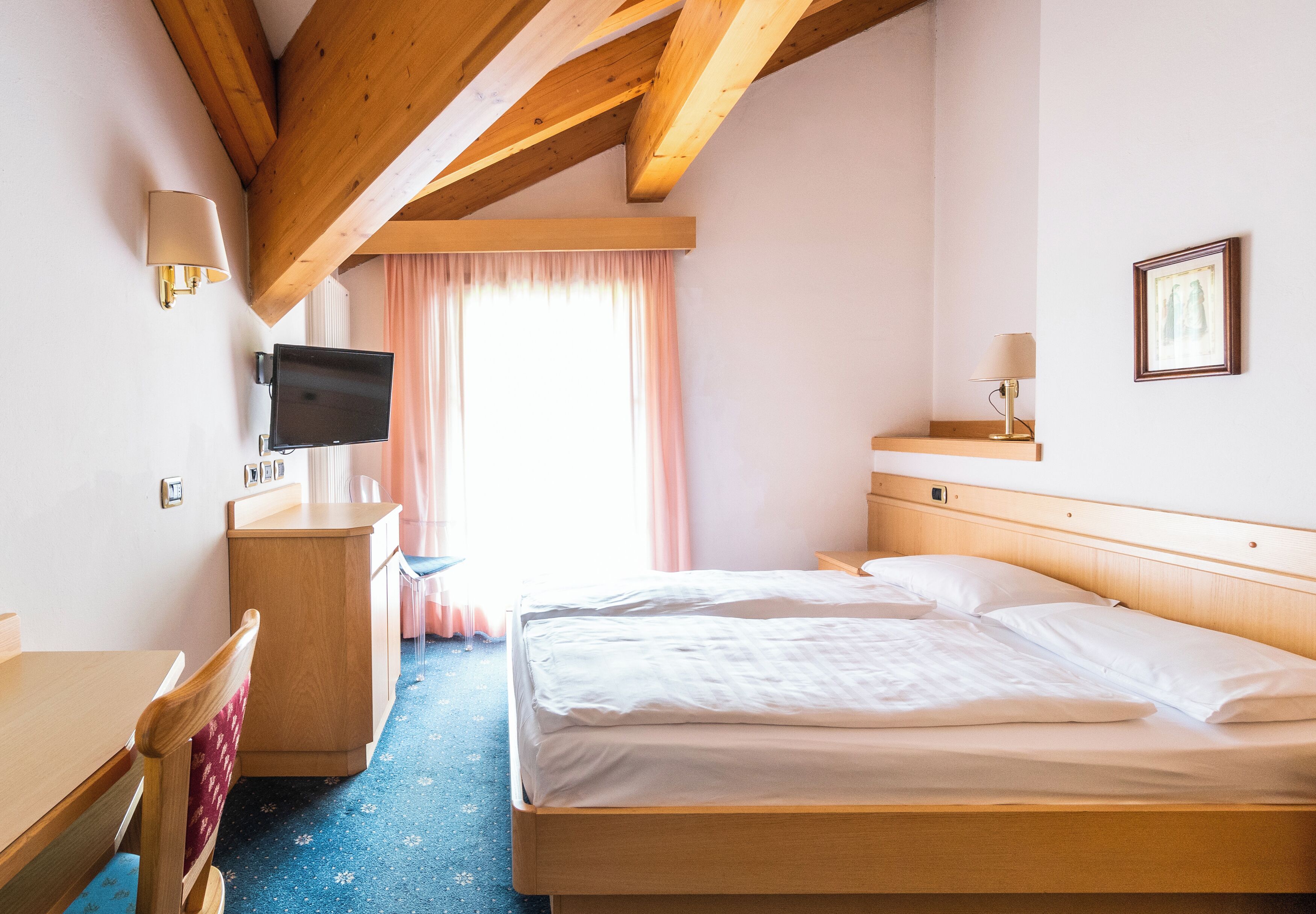 A hotel room with a wooden ceiling, a bed, a desk, a television, and a window with curtains.