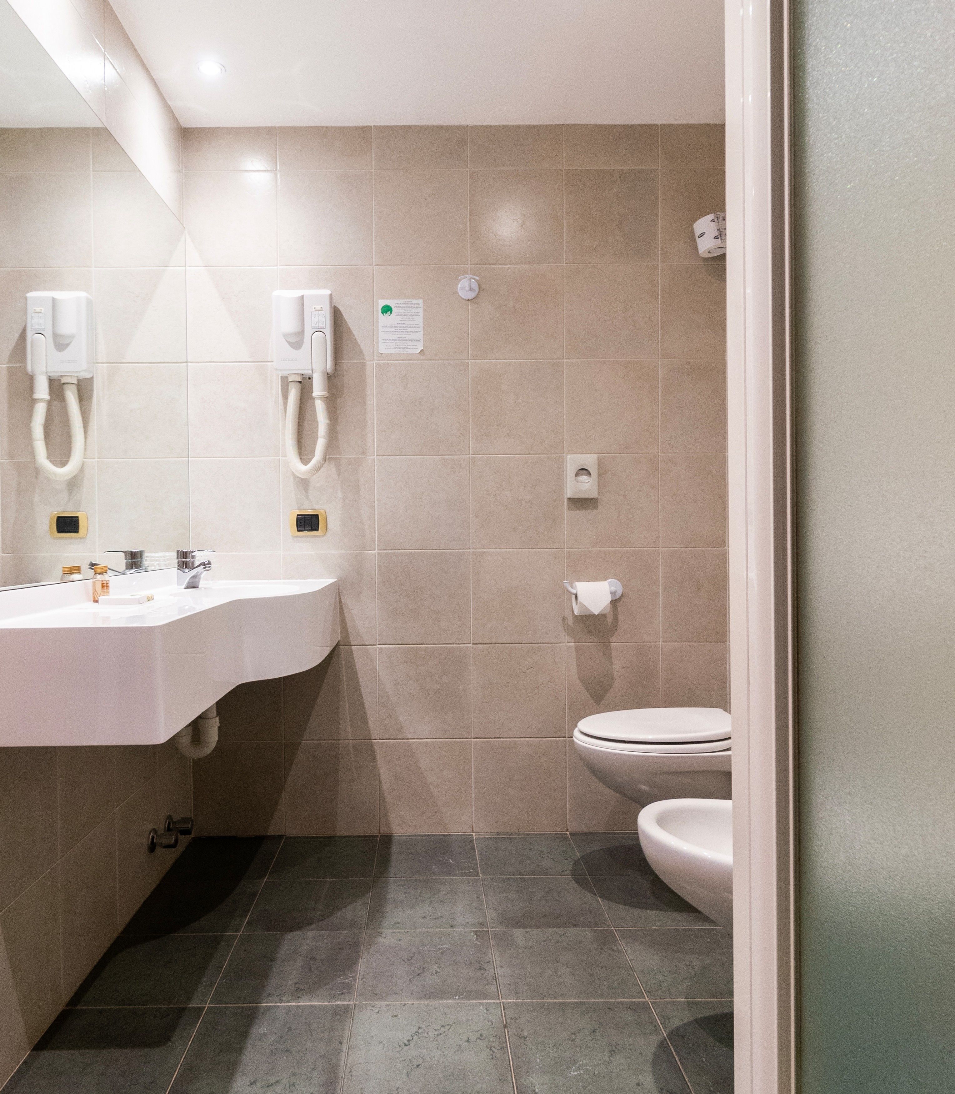 A modern bathroom with a white sink, two towel dispensers, a toilet, and a paper towel dispenser.