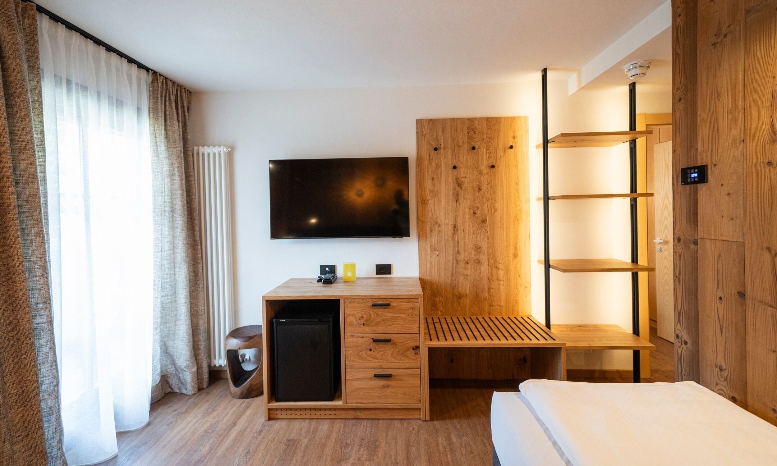 A modern hotel room with a wooden desk, a flat-screen TV, a bed, and a built-in shelf unit.