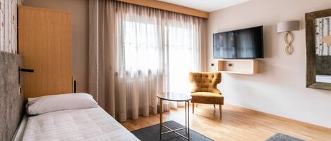 A hotel room with a bed, a small round table, a chair, a TV mounted on the wall, and a window with curtains.
