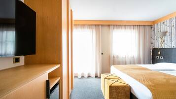 A hotel room with a flat-screen TV, a bed with a headboard, a bedside table, and a window with sheer curtains.