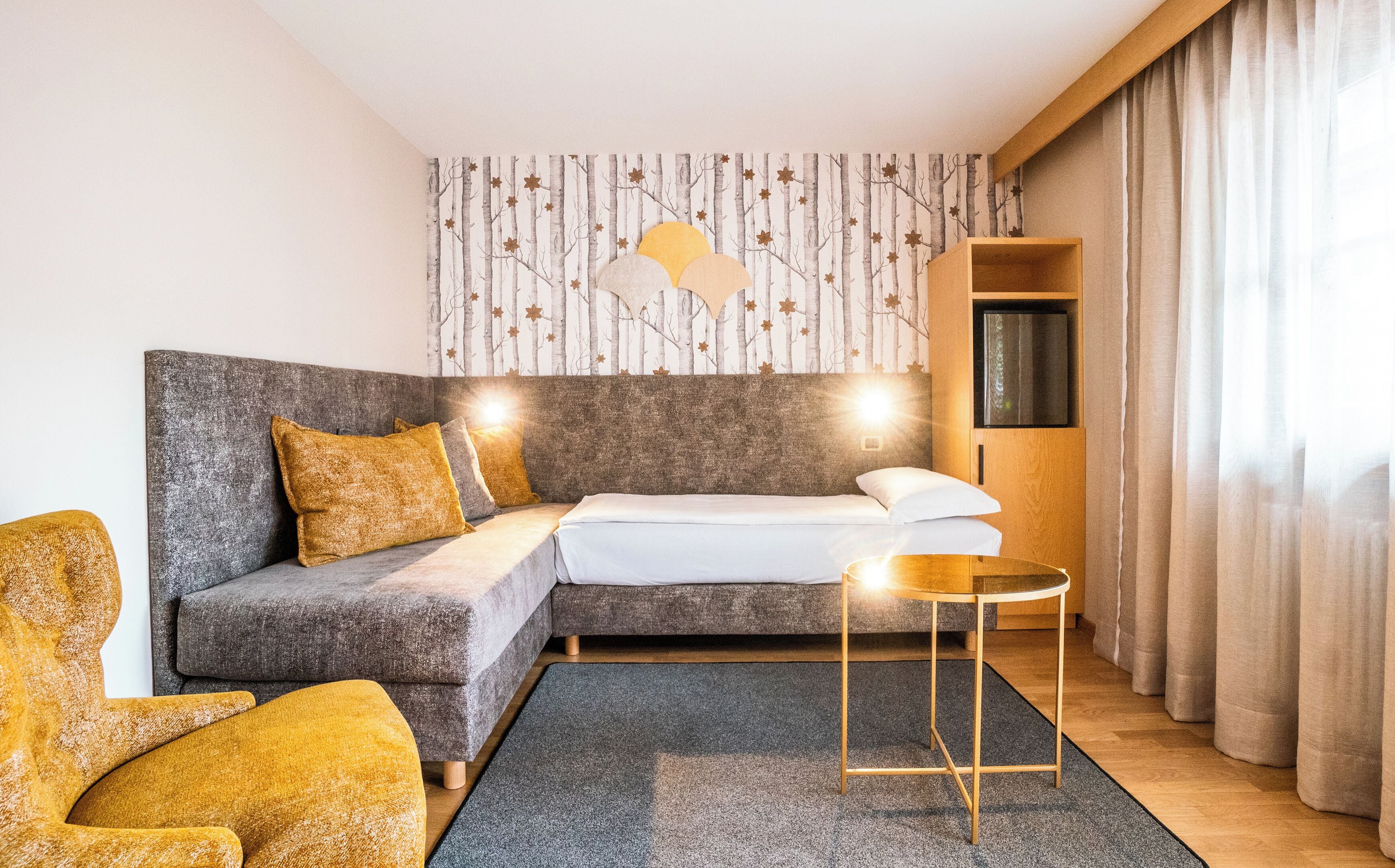 Una habitación de hotel moderna con un sofá gris, una mesita redonda pequeña y una cama con ropa de cama blanca.