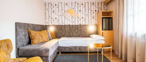 Una habitación de hotel moderna con un sofá gris, una mesita redonda pequeña y una cama con ropa de cama blanca.