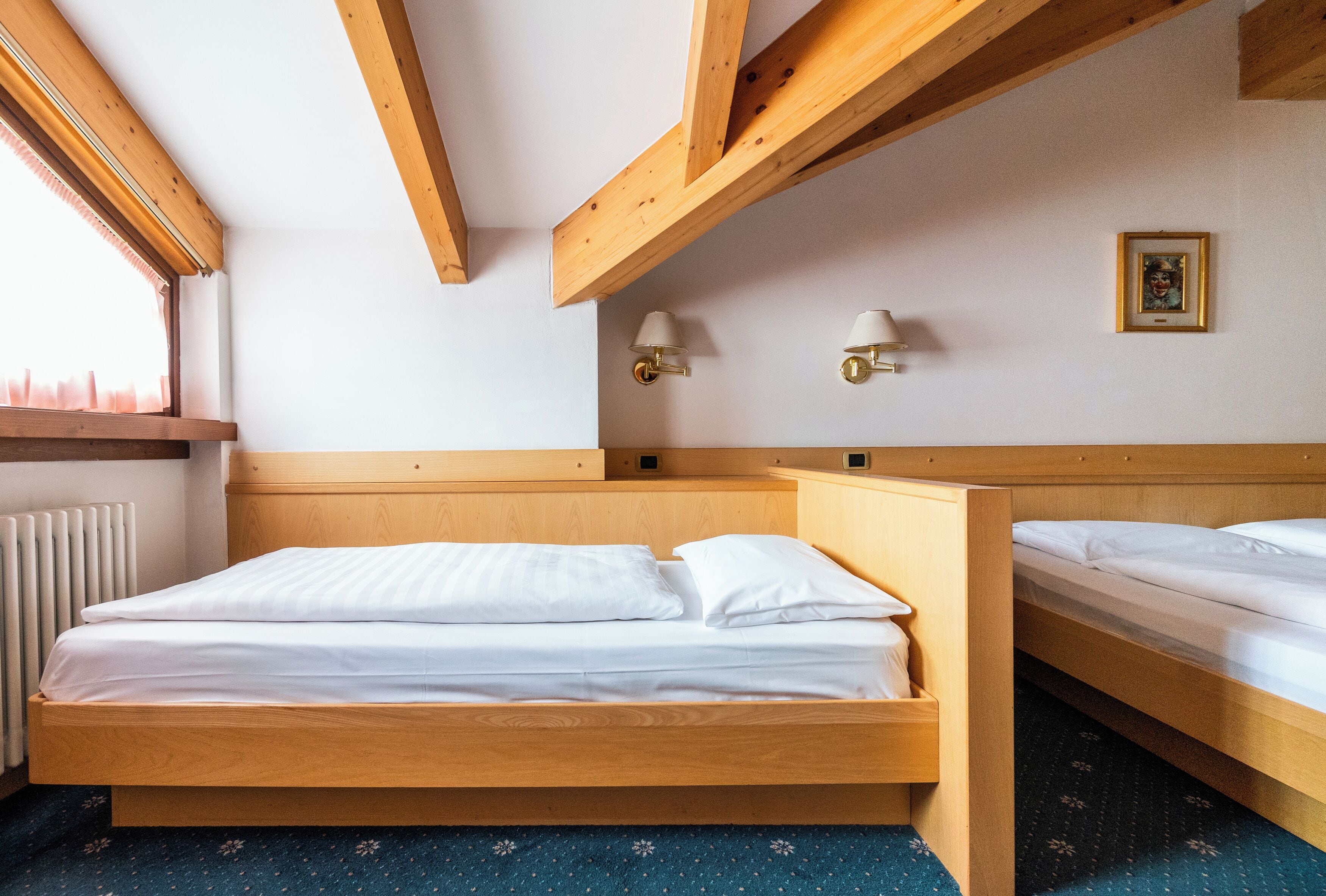 A bedroom with two wooden beds, a window with blinds, a radiator, and two wall-mounted lamps.