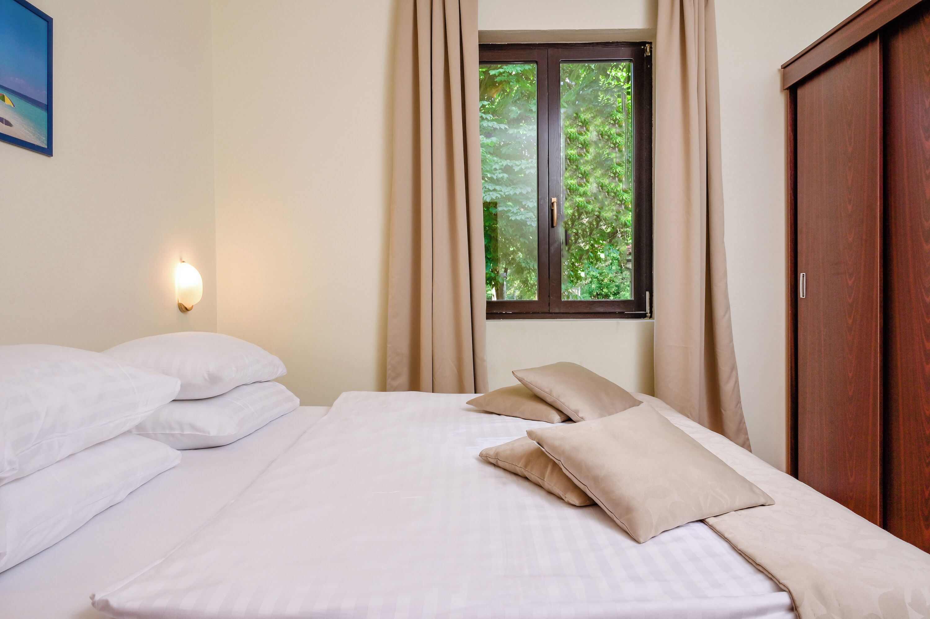 A hotel room with a bed, beige curtains, a window with a view of greenery, and a wooden wardrobe.