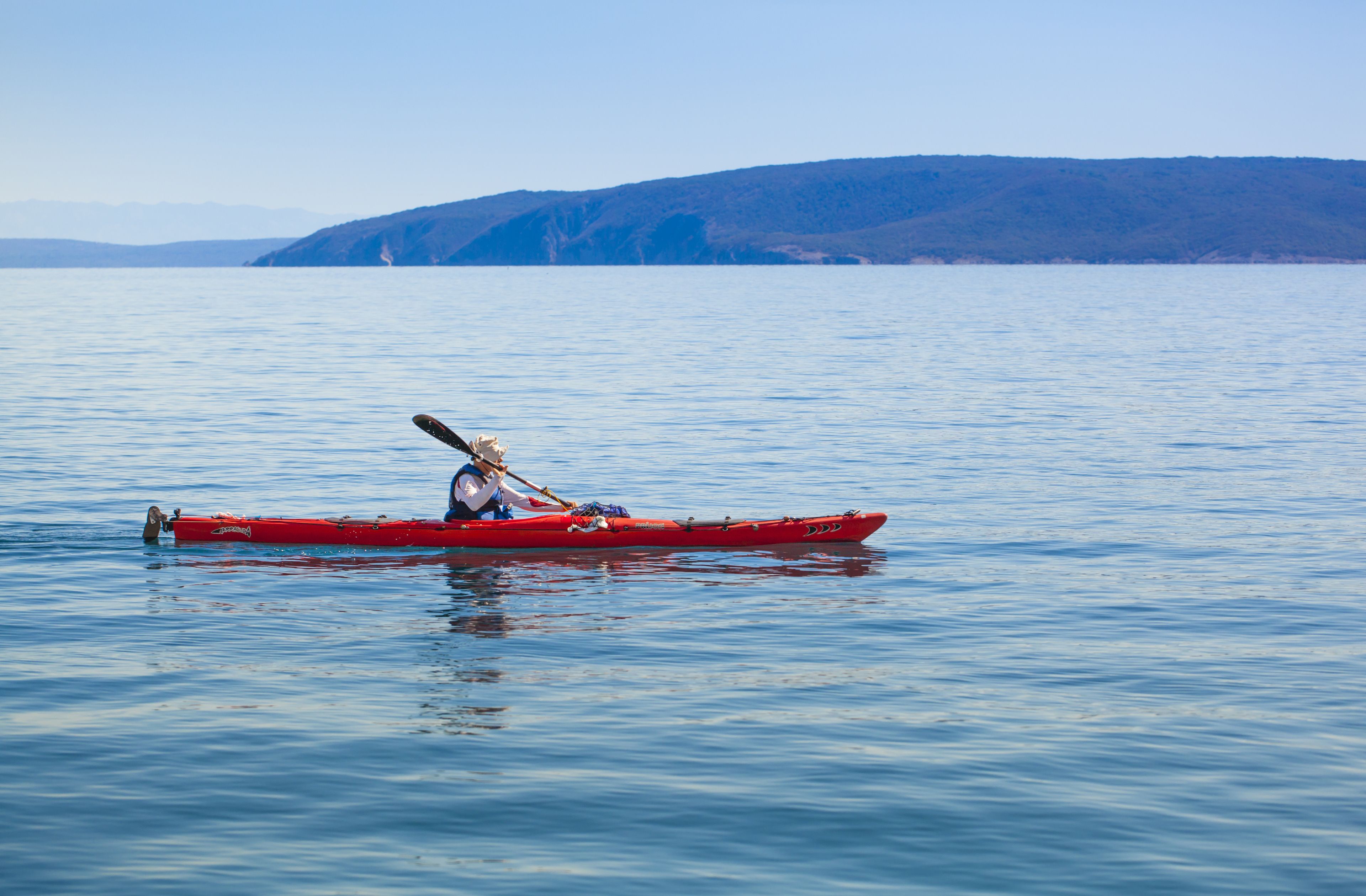 Una persona in kayak rosso su acque calme, con un paesaggio collinare sullo sfondo.