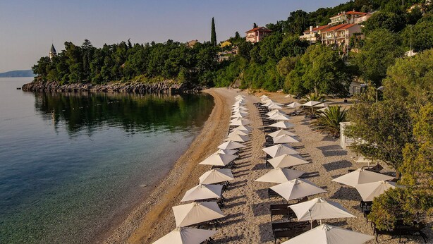 A beach with rows of umbrellas and chairs, a clear view of the water, and buildings on a hillside. - Holiday Resort Medveja - Liburnia (Lovran)