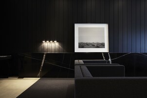 Una habitación moderna con un panel de madera oscura, una fotografía blanco y negro enmarcada, y una zona de estar con un sofá oscuro y una mesita.