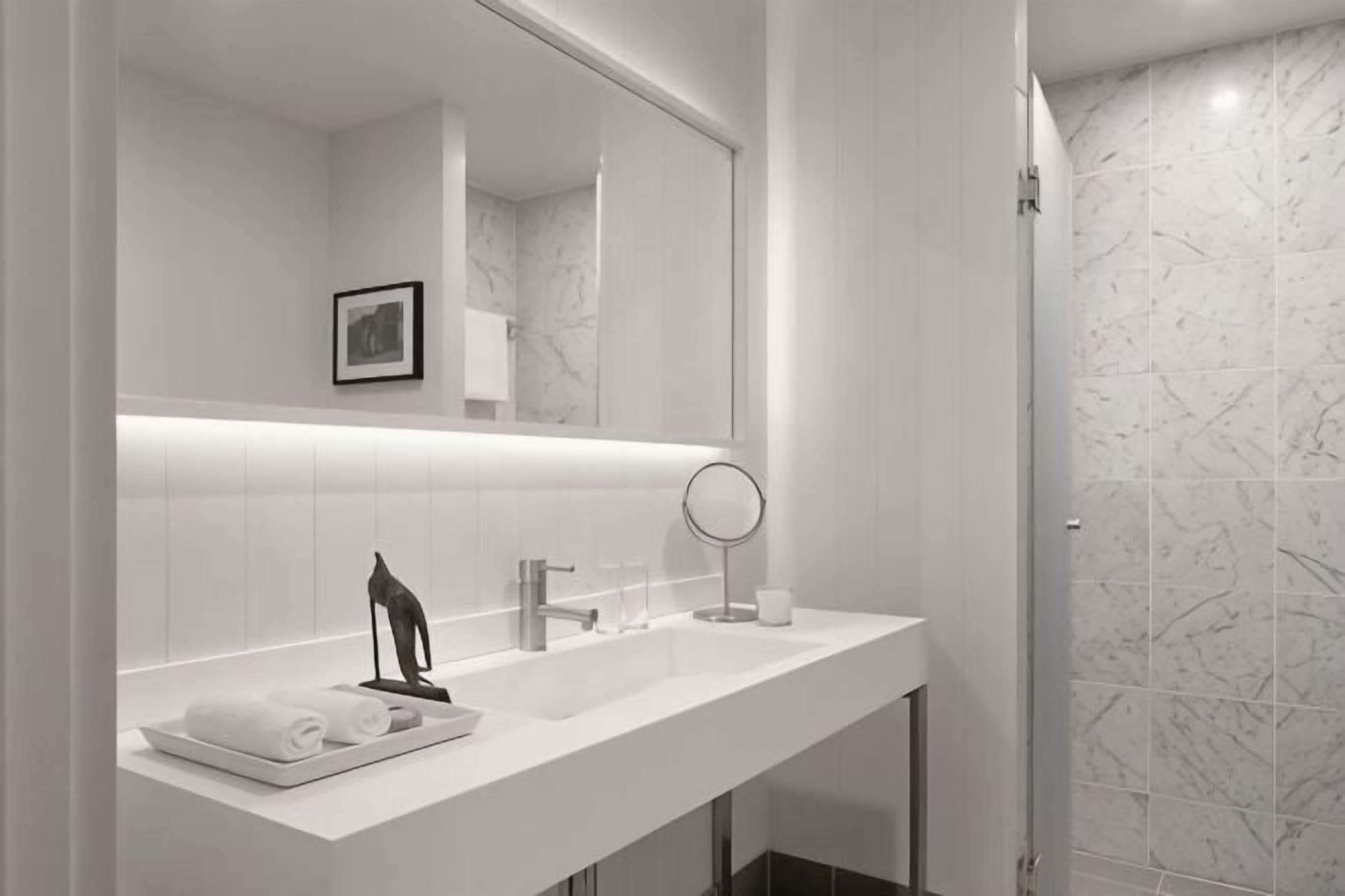 Un baño moderno con un tocador blanco, un espejo grande y una zona de ducha con azulejos de mármol.