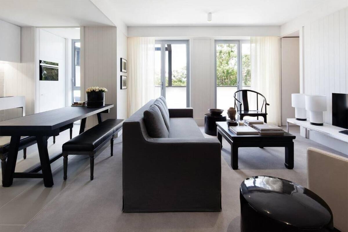 A modern living room with a black sofa, a dining table, and a black chair. There is a window with curtains, a television, and a lamp.